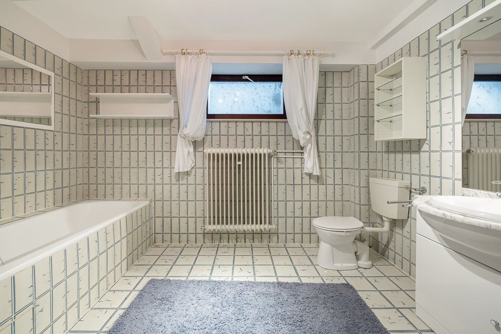 Helles Badezimmer mit Badewanne, WC, Waschbecken, Regal und Fenster mit Vorhängen.
