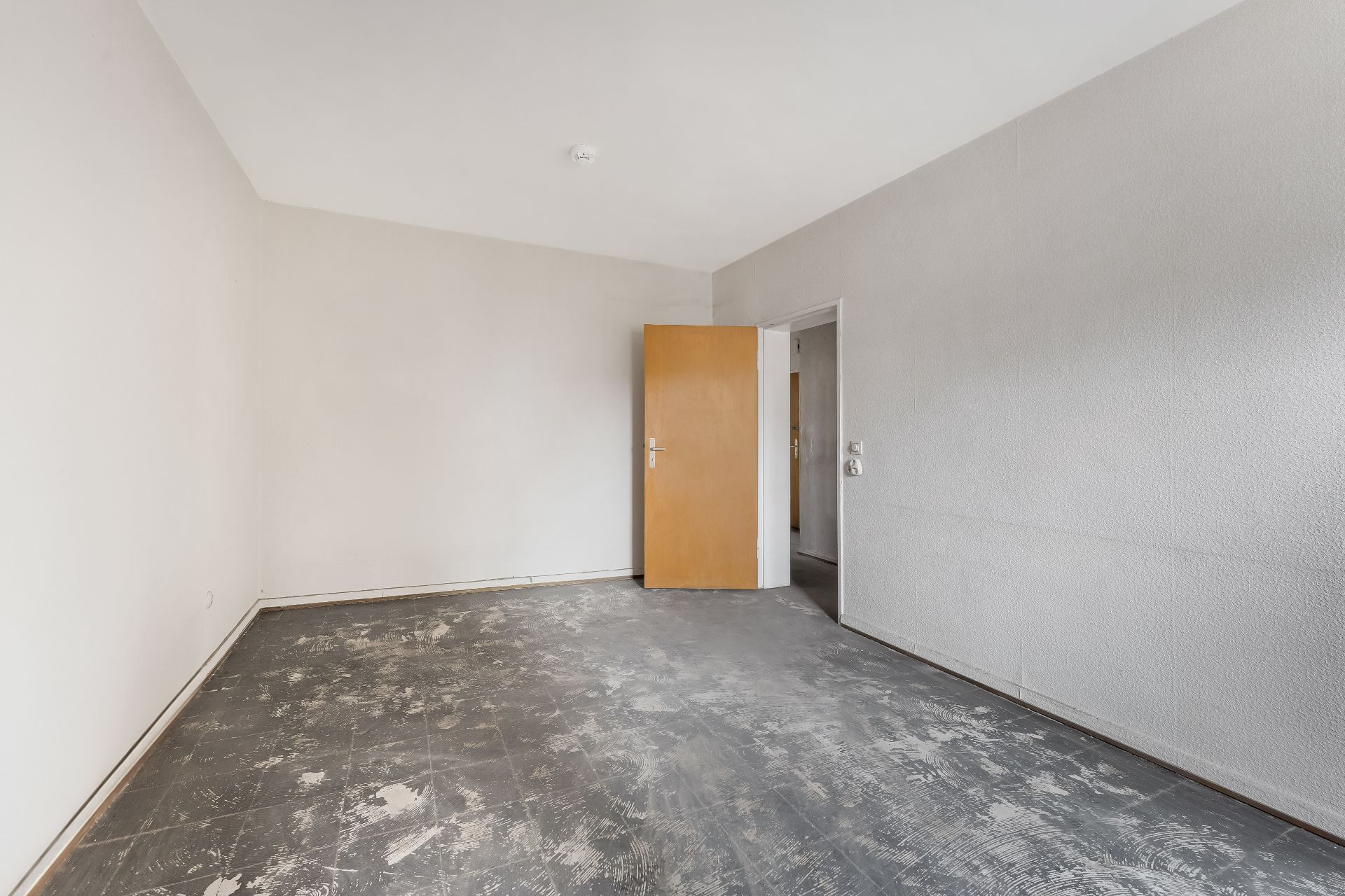 Leerer Raum mit grauem Boden, weißen Wänden und geöffneter Holztür.