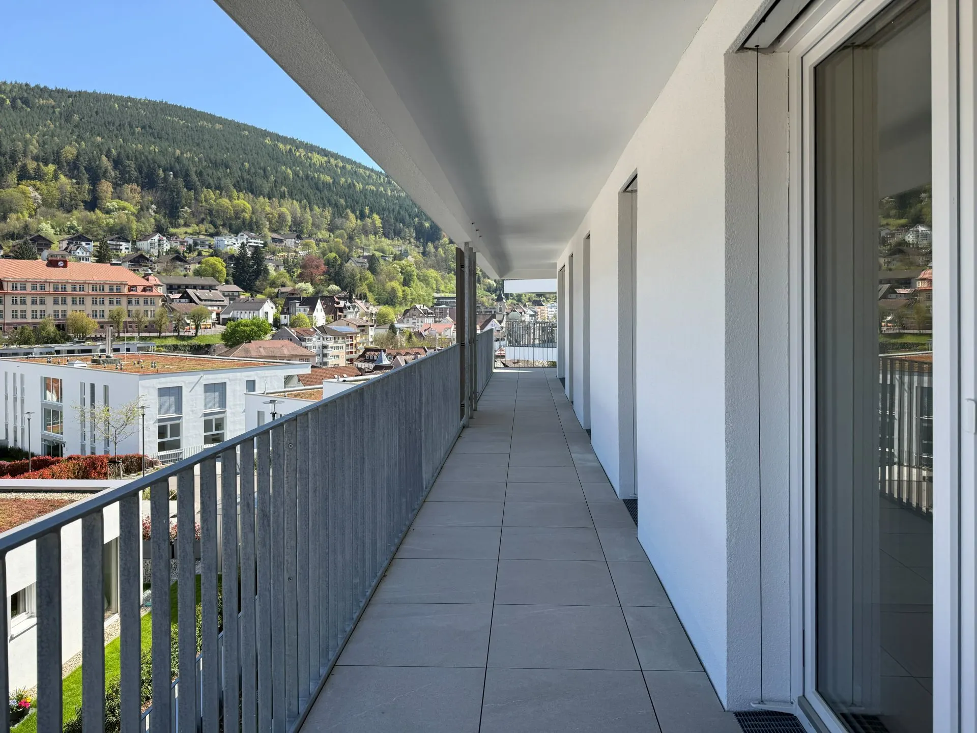 Langer Balkon mit Geländer an einem Wohnhaus, Blick auf Häuser und bewaldete Hügel.