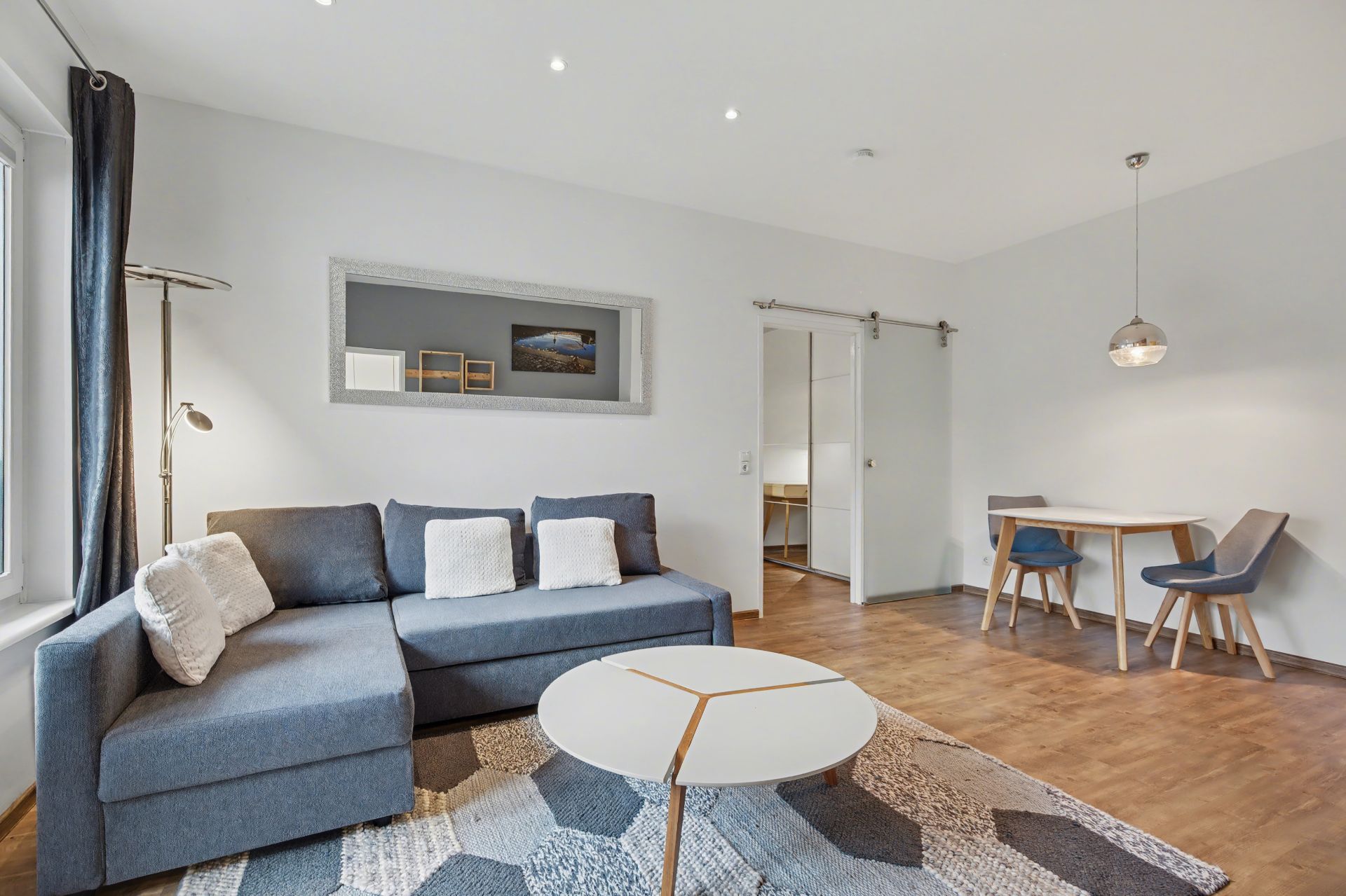 Modernes Wohnzimmer mit grauem Sofa, rundem Couchtisch, Esstisch und zwei Stühlen auf Holzfußboden.