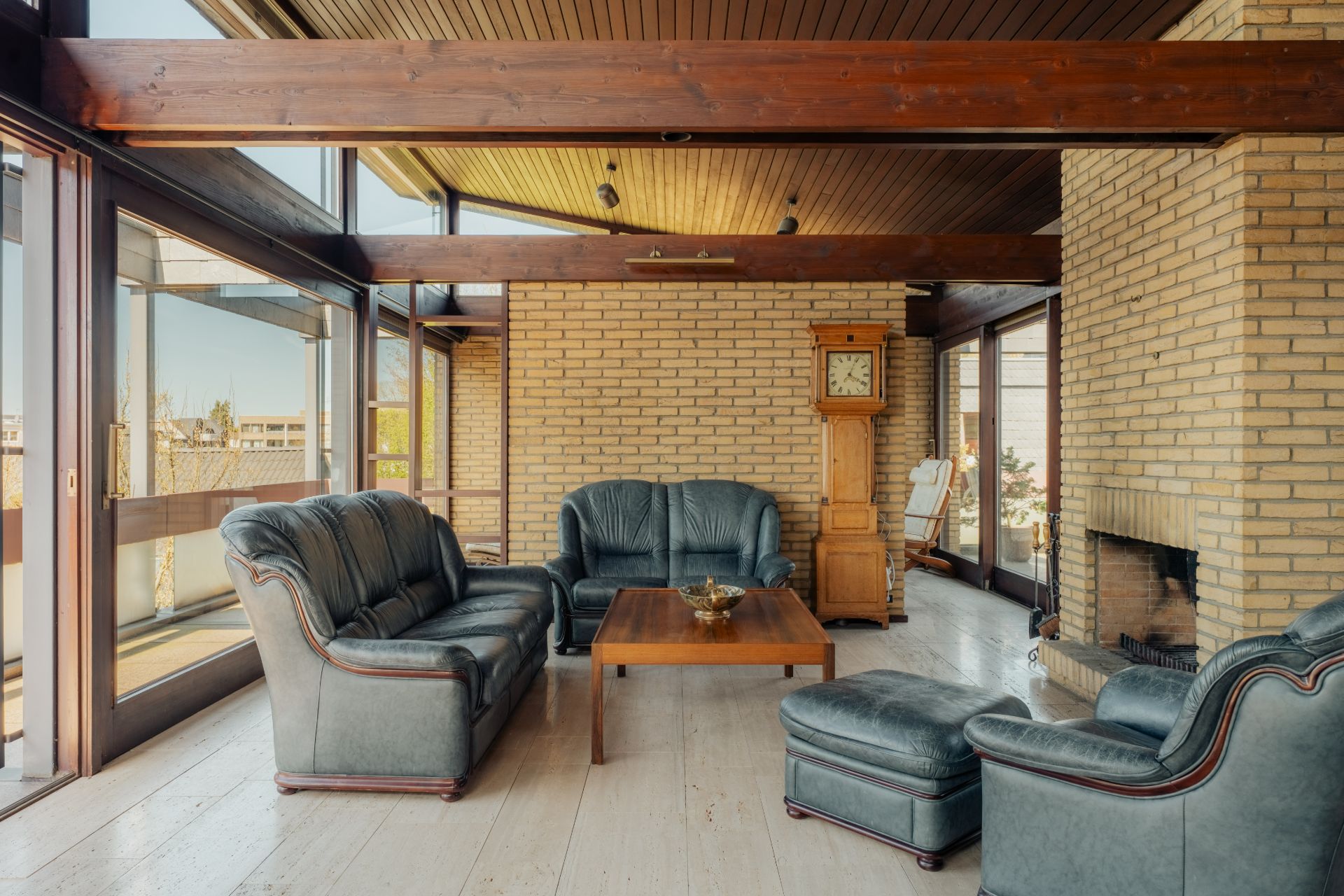 Wohnzimmer mit dunklen Ledersofas, Holztisch, Kamin und Standuhr vor einer Backsteinwand.