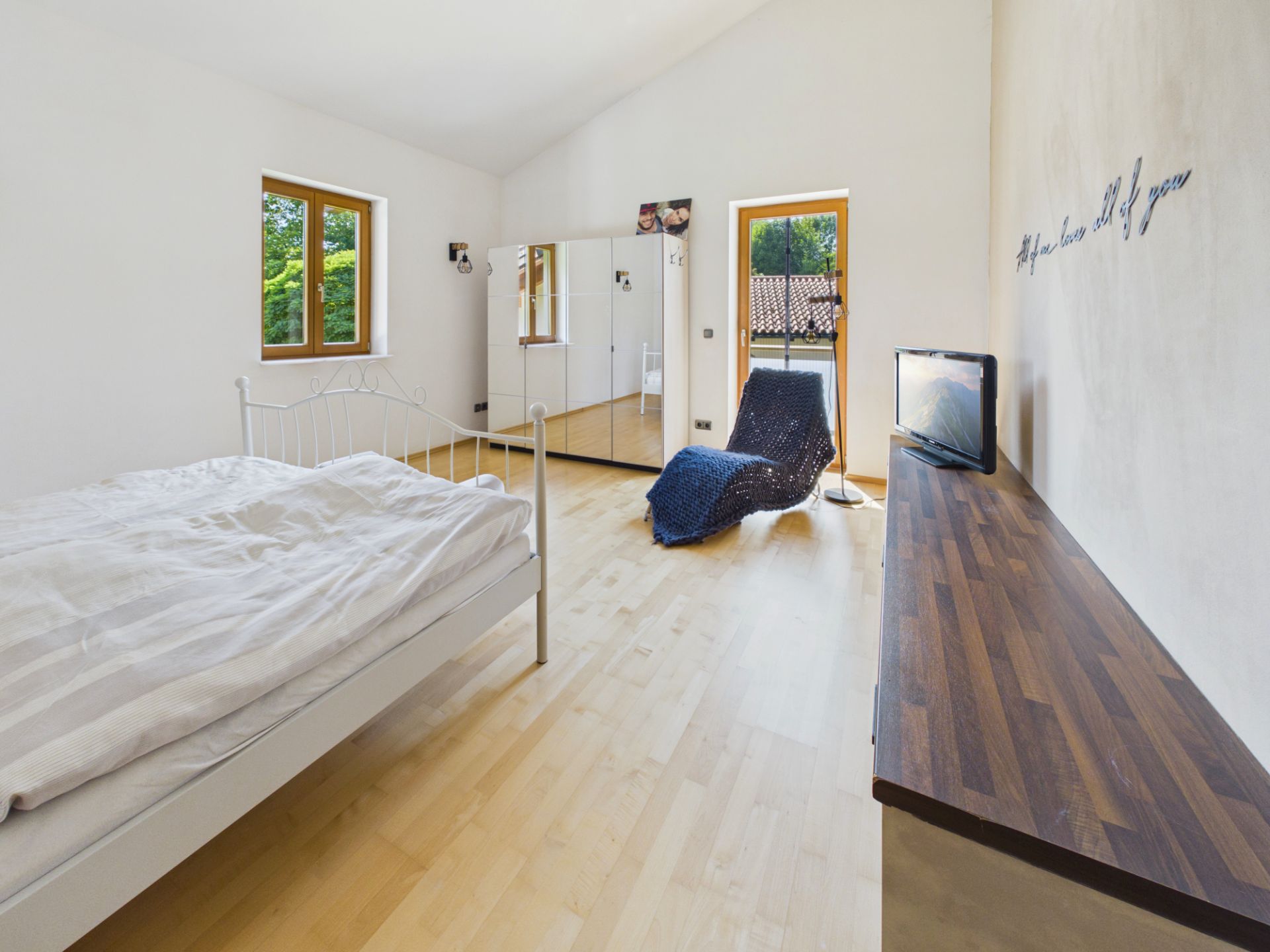 Helles Schlafzimmer mit Doppelbett, Fernseher, Sessel, Kommode und großem Spiegel.