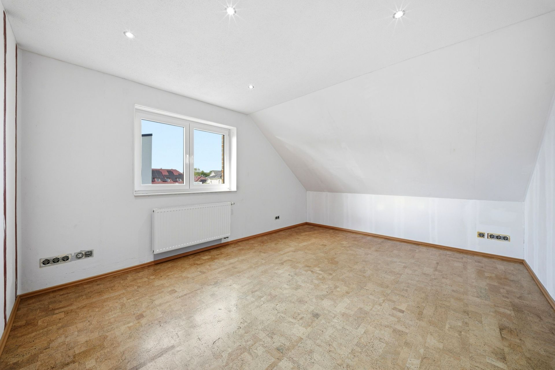 Helles, leeres Zimmer mit weißer Wand, Fenster, Heizkörper und schräger Decke.