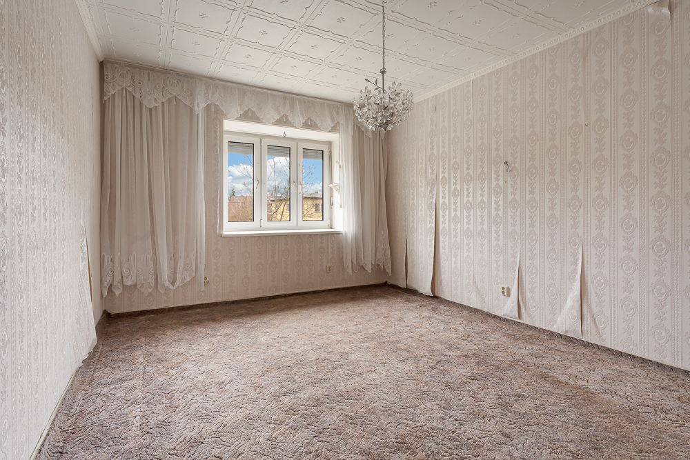 Leeres Zimmer mit Teppichboden, Tapeten, Fenster mit Vorhängen und Deckenleuchte.