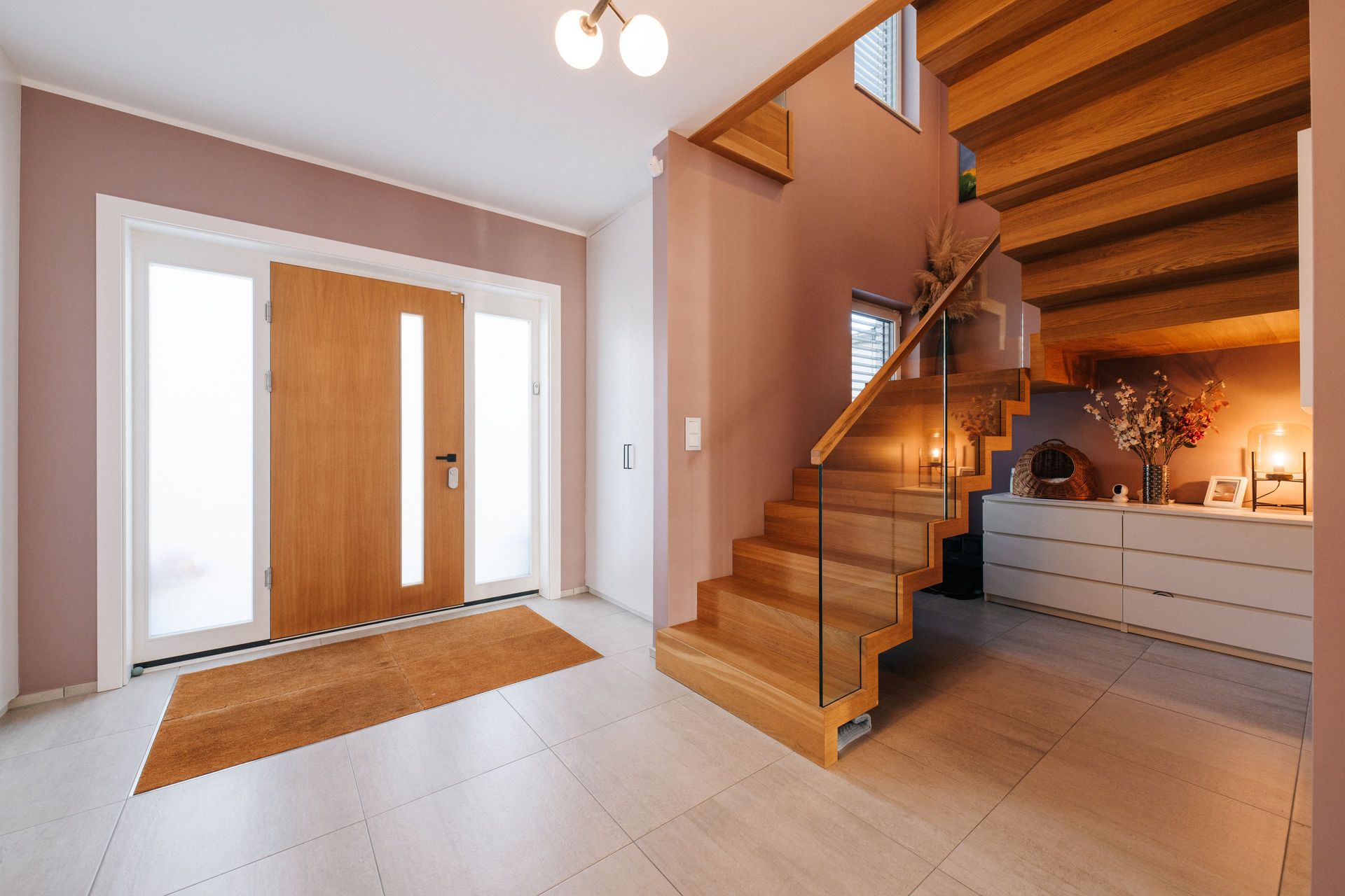 Eingangsbereich mit Holztür, Treppe aus Holz und Glasgeländer, Kommode mit Dekoration.