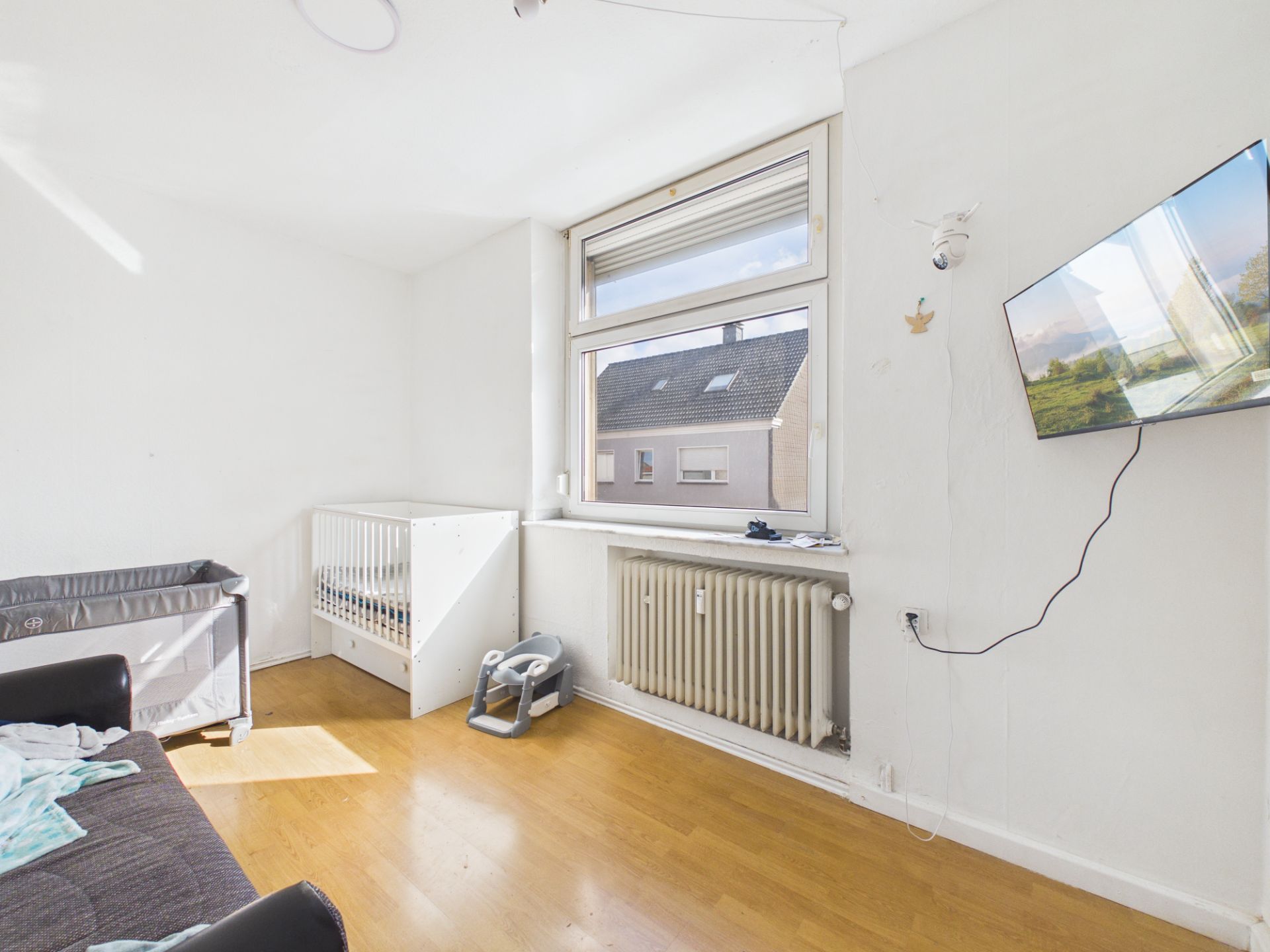 Helles Zimmer mit Gitterbett, Sofa, Fenster und an der Wand montiertem Fernseher.
