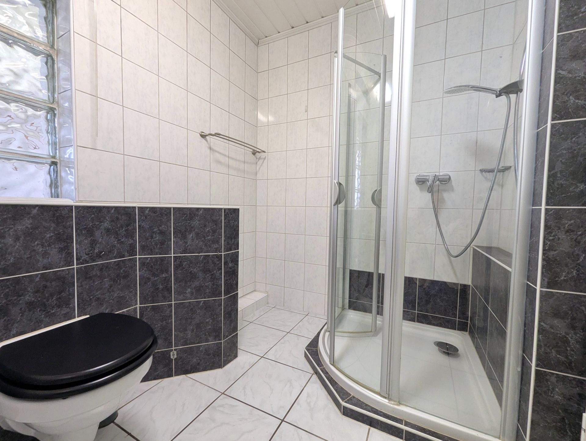 Gefliestes Badezimmer mit Dusche, WC mit schwarzem Sitz und Handtuchhalter.