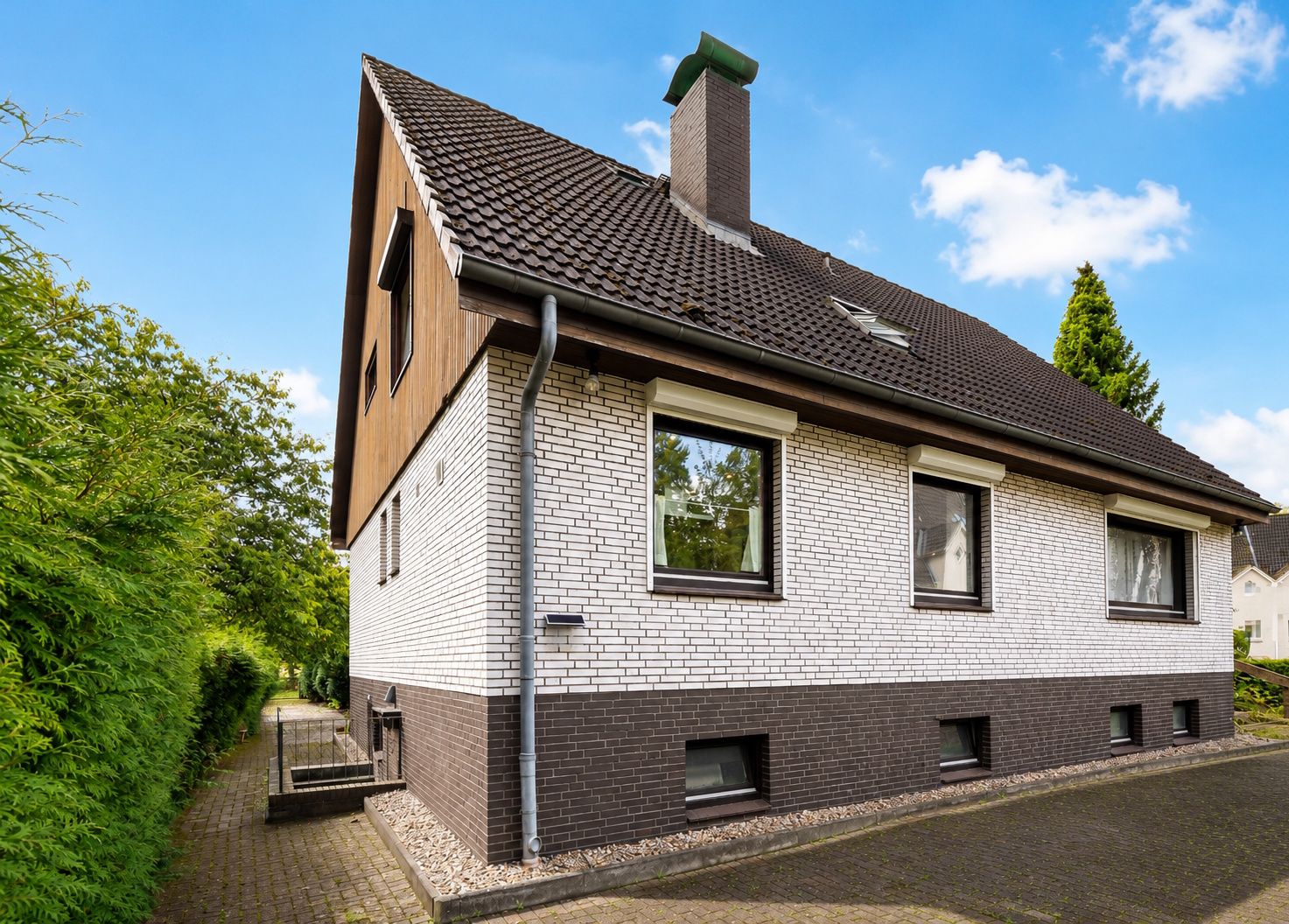 Zweistöckiges Wohnhaus mit dunklem Dach, weißen und braunen Klinkersteinen, großen Fenstern und Schornstein.