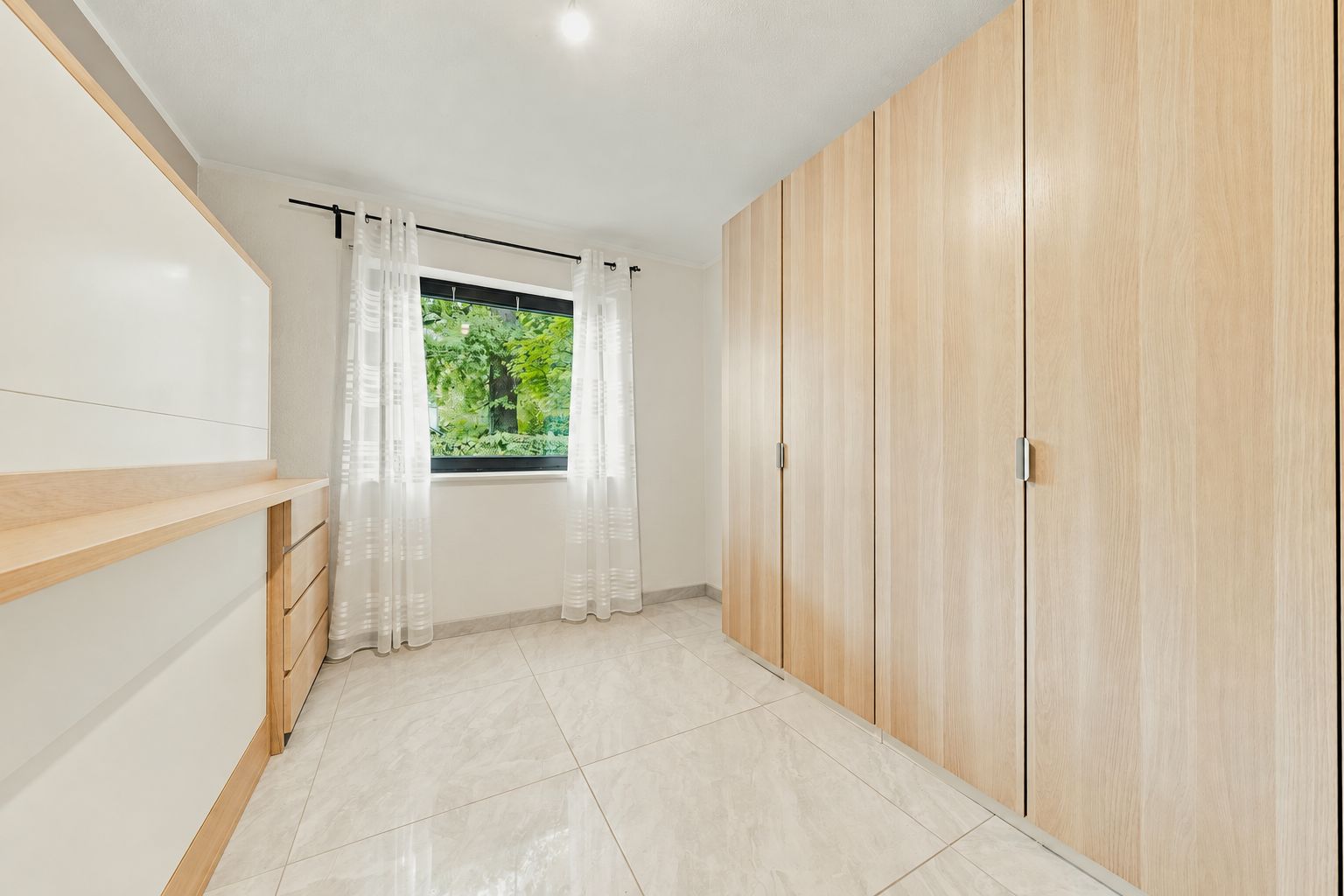 Heller Raum mit großem Holzkleiderschrank, Kommode und Fenster mit weißen Gardinen.