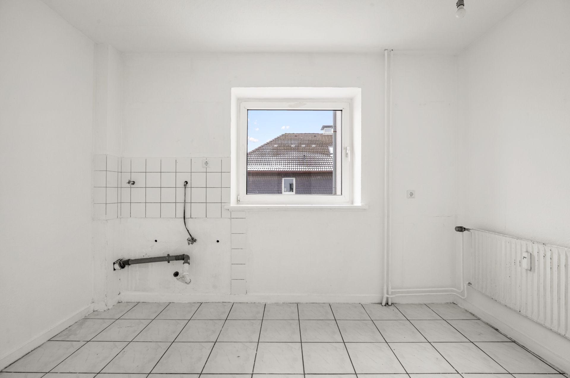 Leerer, weiß gefliester Raum mit Fenster, sichtbaren Rohren und Heizkörper.