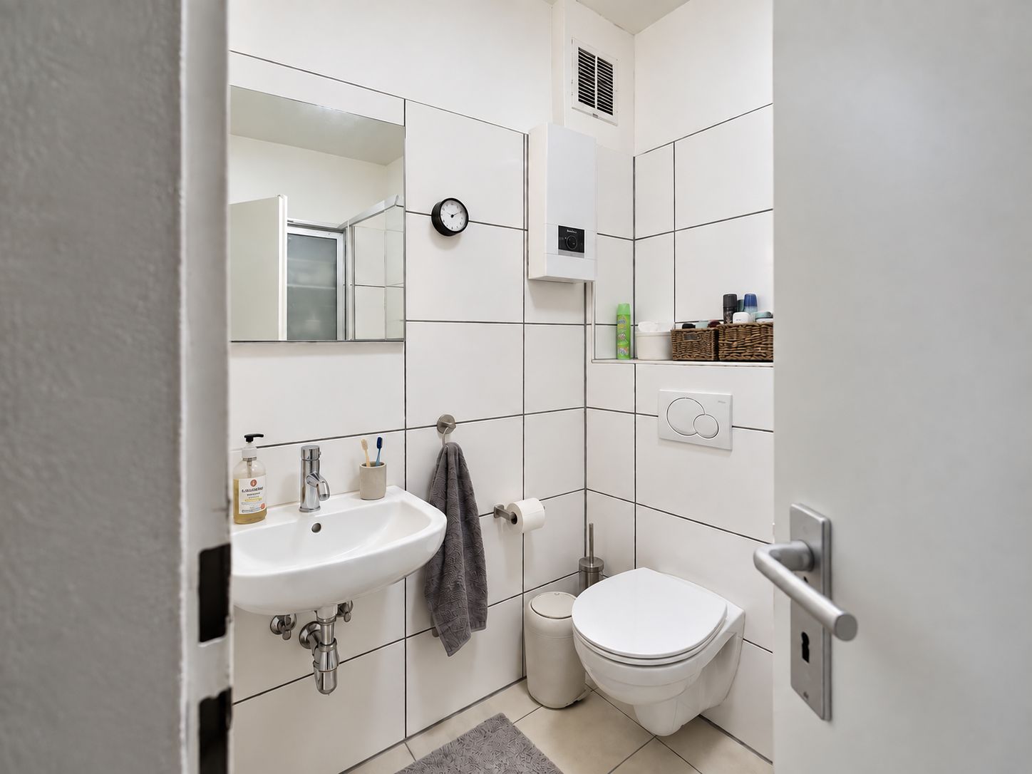 Kleines Badezimmer mit Waschbecken, Wandtoilette, Spiegel und weißen Fliesen.