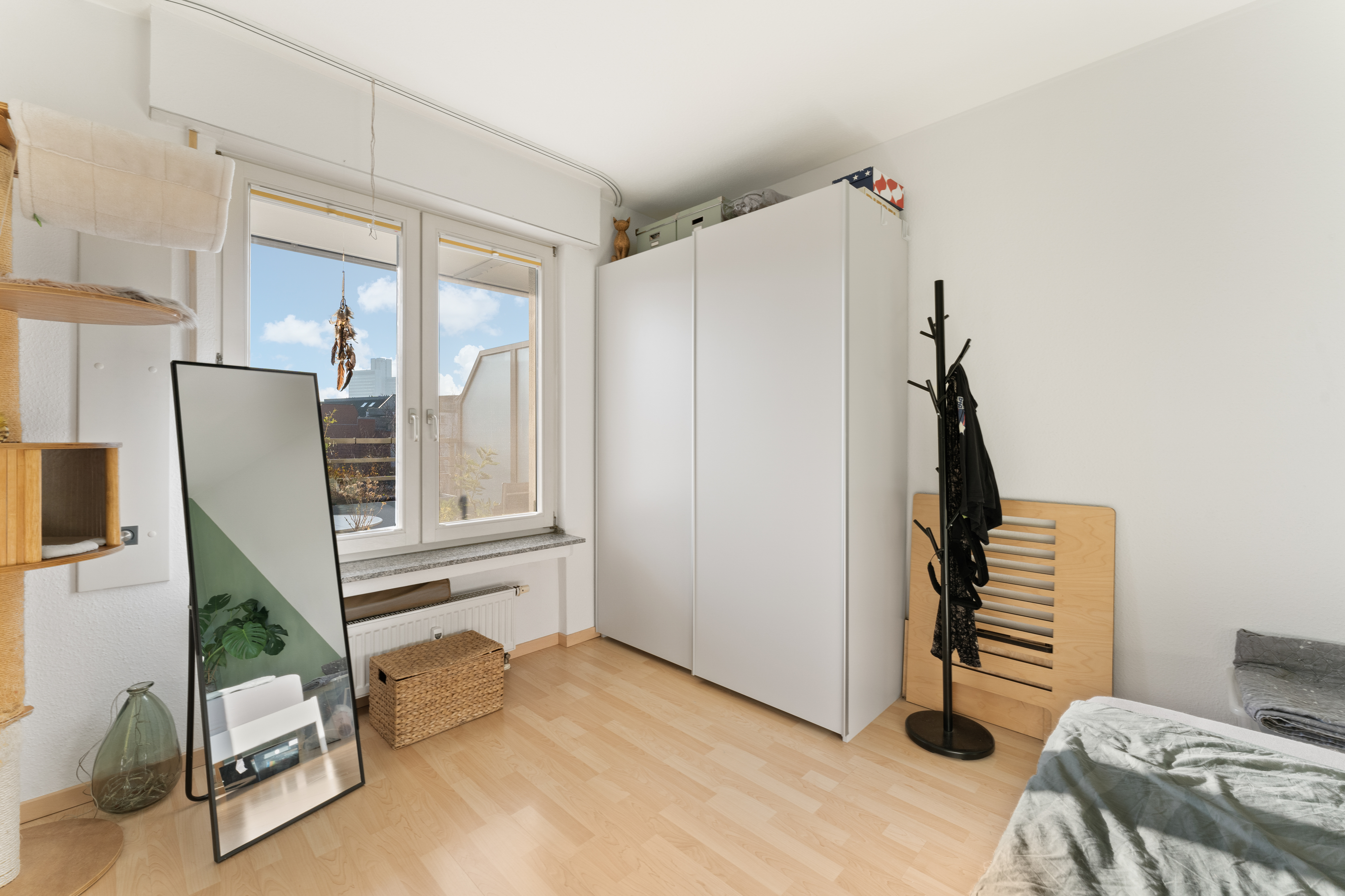 Helles Schlafzimmer mit großem Fenster, weißem Schrank, Spiegel und Garderobenständer.