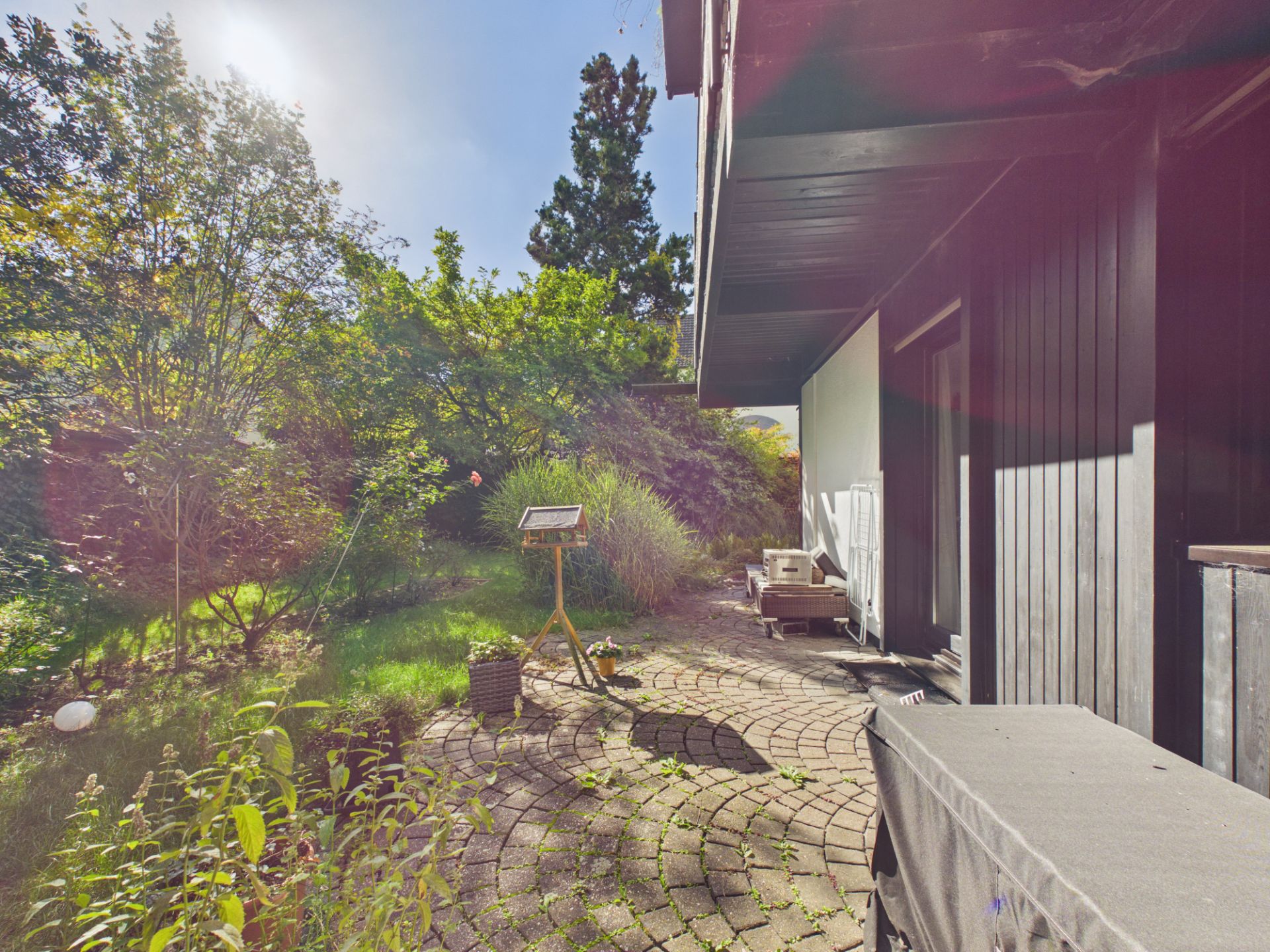 Gepflasterte Terrasse mit Vogelhaus und Garten, seitlich an einem Wohnhaus, sonnig und grün.