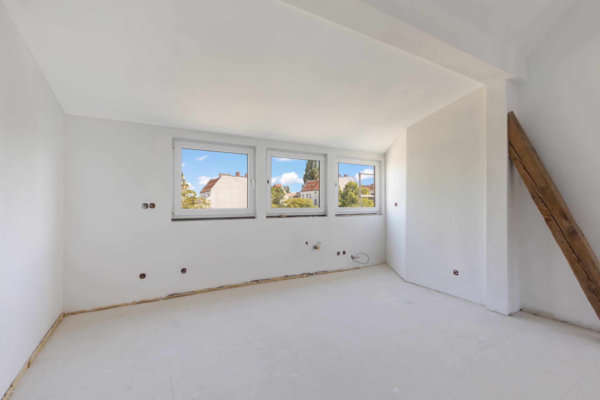 Heller leerer Raum mit drei Fenstern und schräger Decke, teils sichtbarem Holzbalken.