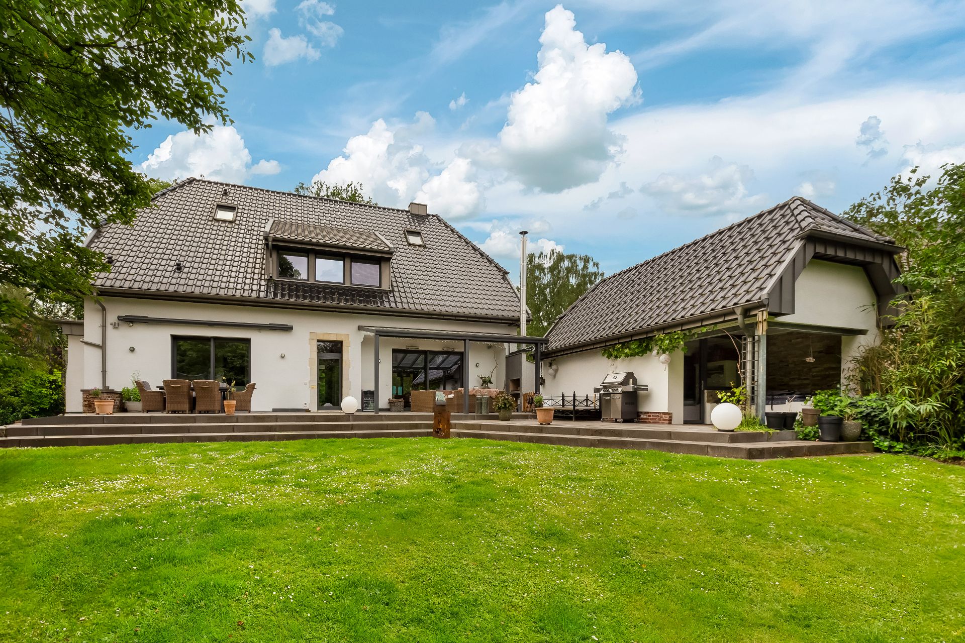Großes Einfamilienhaus mit Terrasse, Gartenmöbeln und gepflegtem Rasen im Vordergrund.