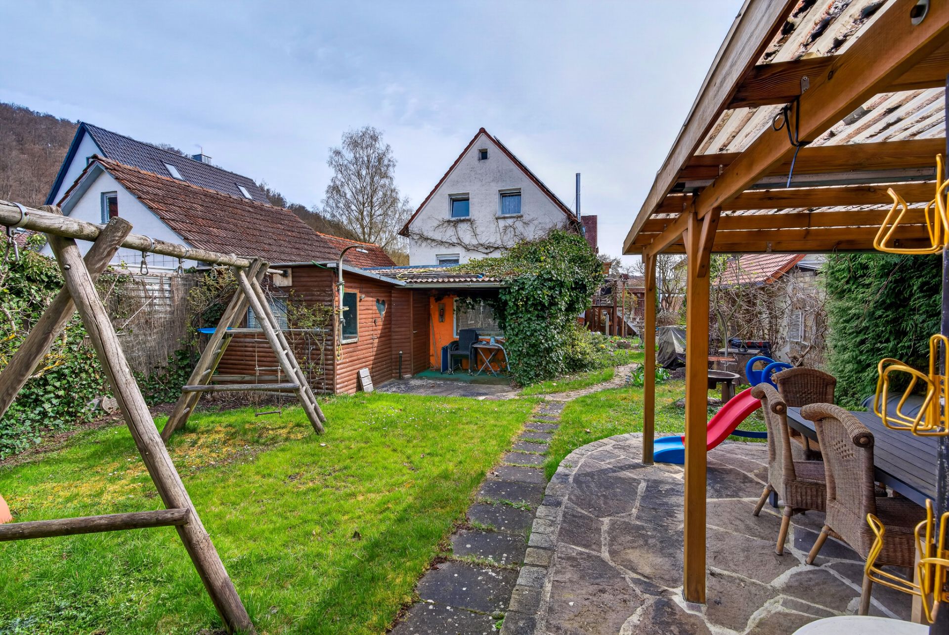 Garten mit Schaukel, Rutsche, Holzschuppen und überdachter Terrasse mit Sitzbereich.