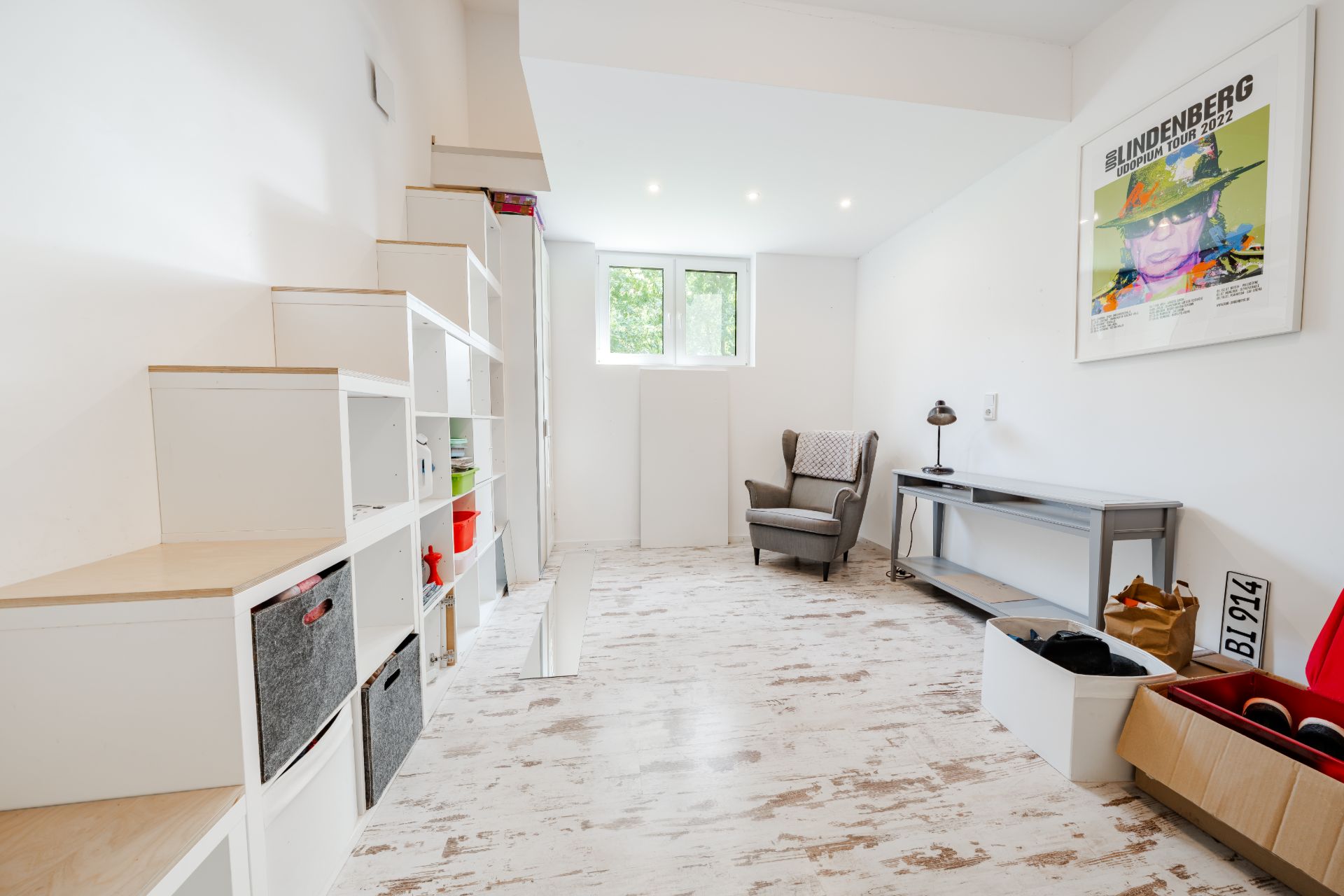 Helles Zimmer mit Regal, Sessel, Schreibtisch und Wandbild, Fenster im Hintergrund.