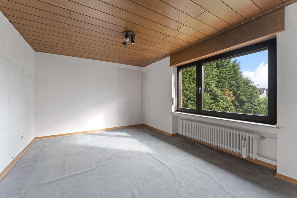 Leeres Zimmer mit Holzdecke, großem Fenster und Heizkörper, Blick auf grüne Bäume.