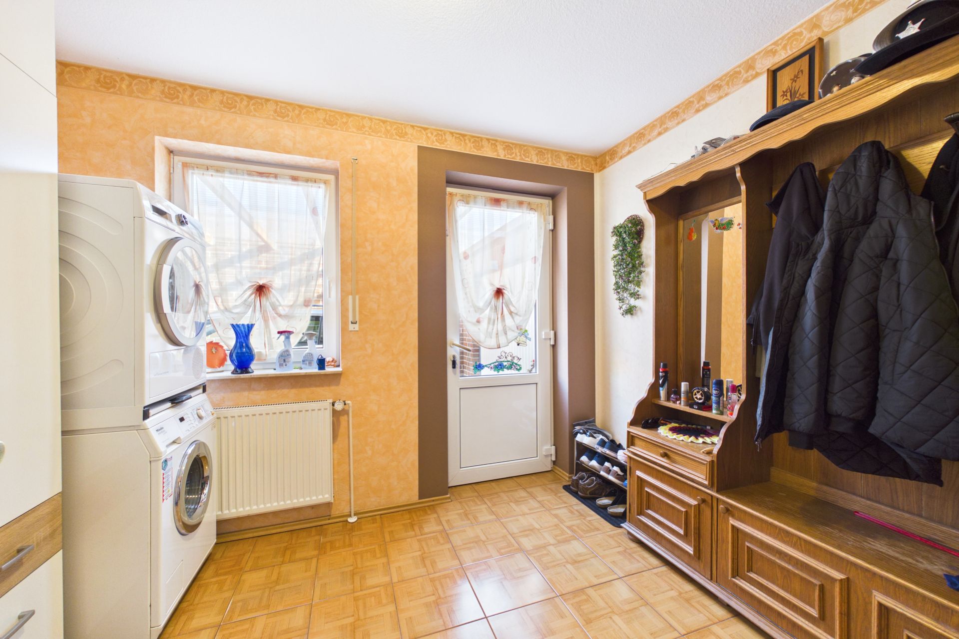 Hausflur mit Garderobe, Schuhregal, Waschmaschine, Trockner und zwei Fenstern mit Vorhängen.