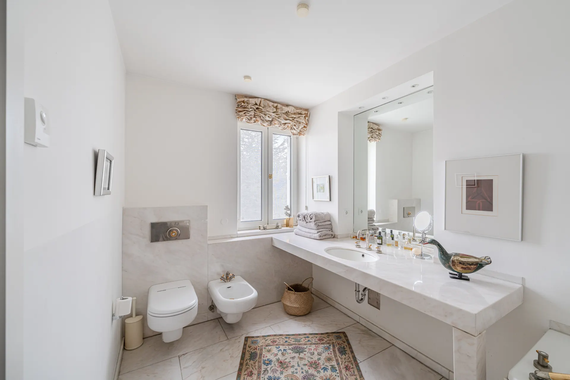 Helles Badezimmer mit Toilette, Bidet, großem Spiegel und Fenster, dekoriert mit Handtüchern und Teppich.