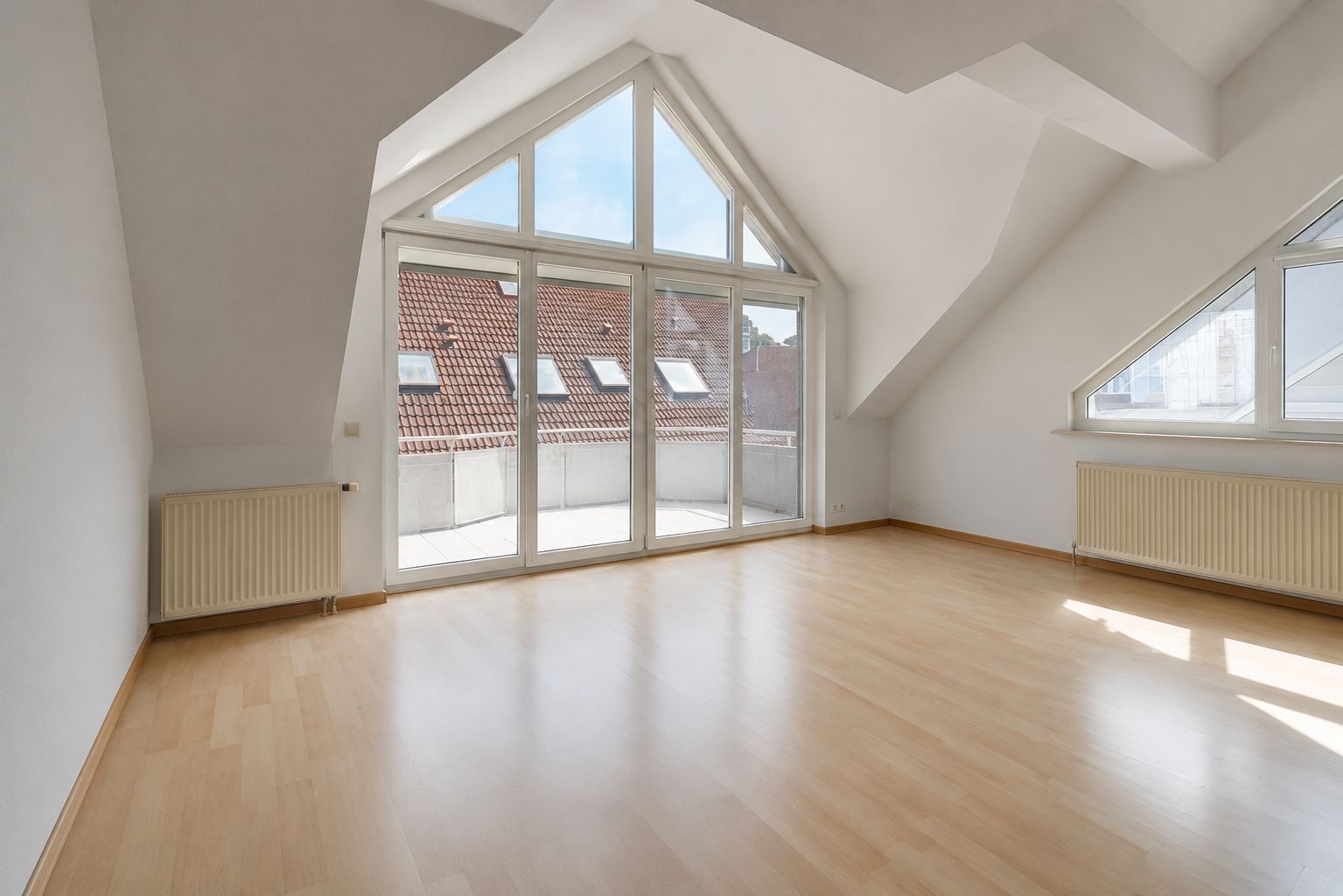 Heller Raum mit Dachschrägen, großem Fenster, Parkettboden und Zugang zu einem Balkon.