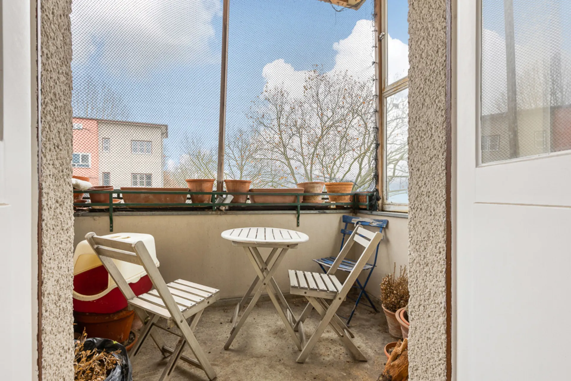 Kleiner Balkon mit drei Stühlen, rundem Tisch und Blumentöpfen am Geländer.