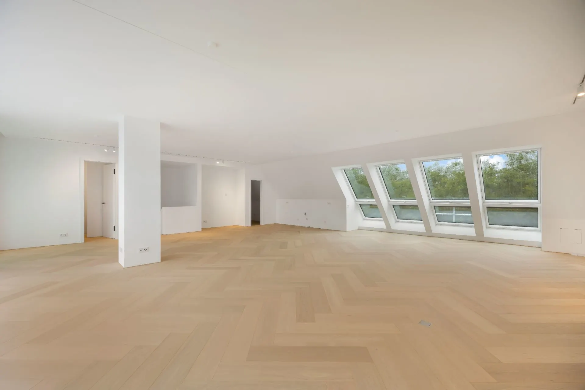 Helles, leeres Zimmer mit Holzfußboden und großen, schrägen Fenstern an einer Wand.