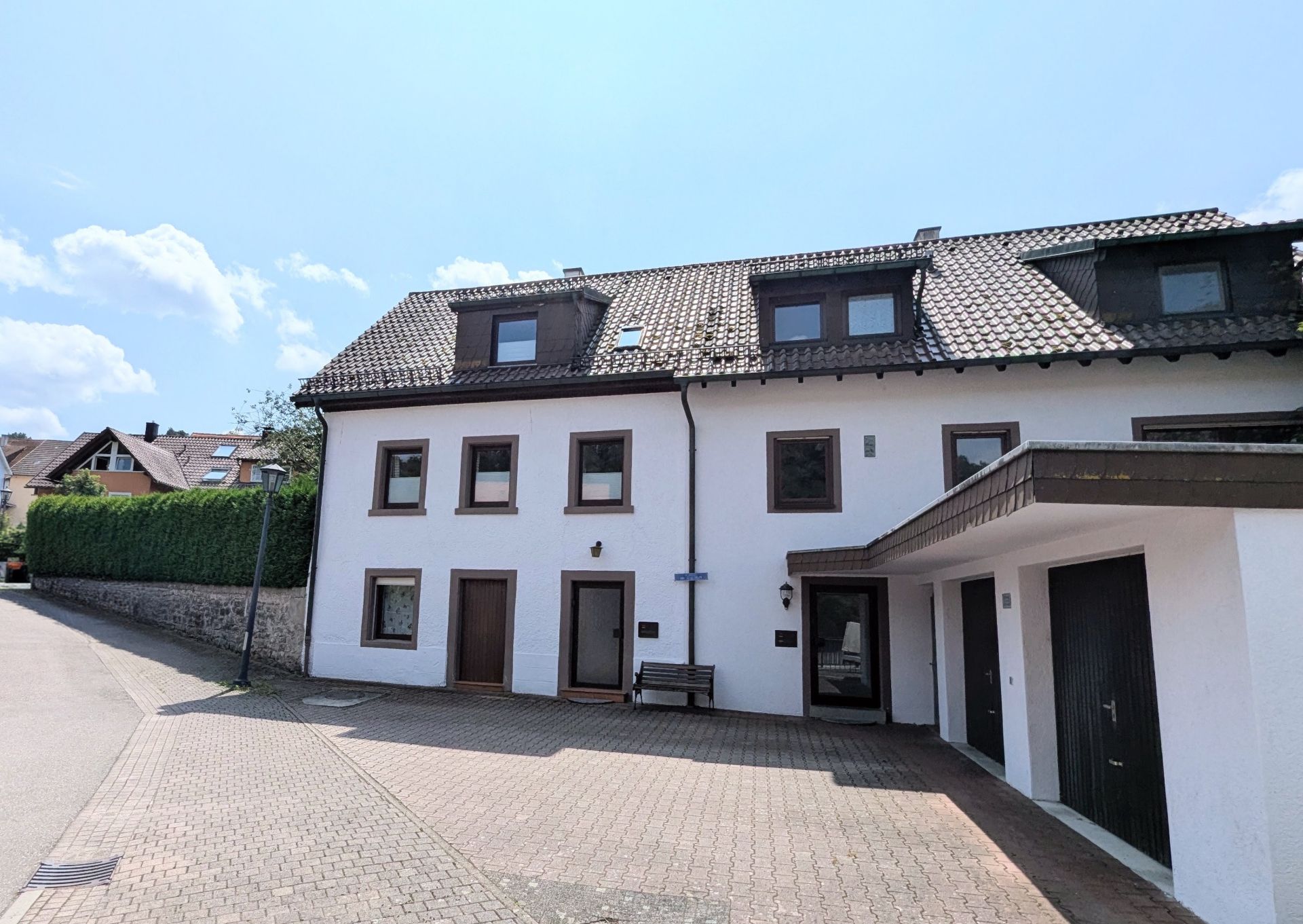 Zweistöckiges Wohnhaus mit dunklem Dach und Garage, davor gepflasterter Hof.