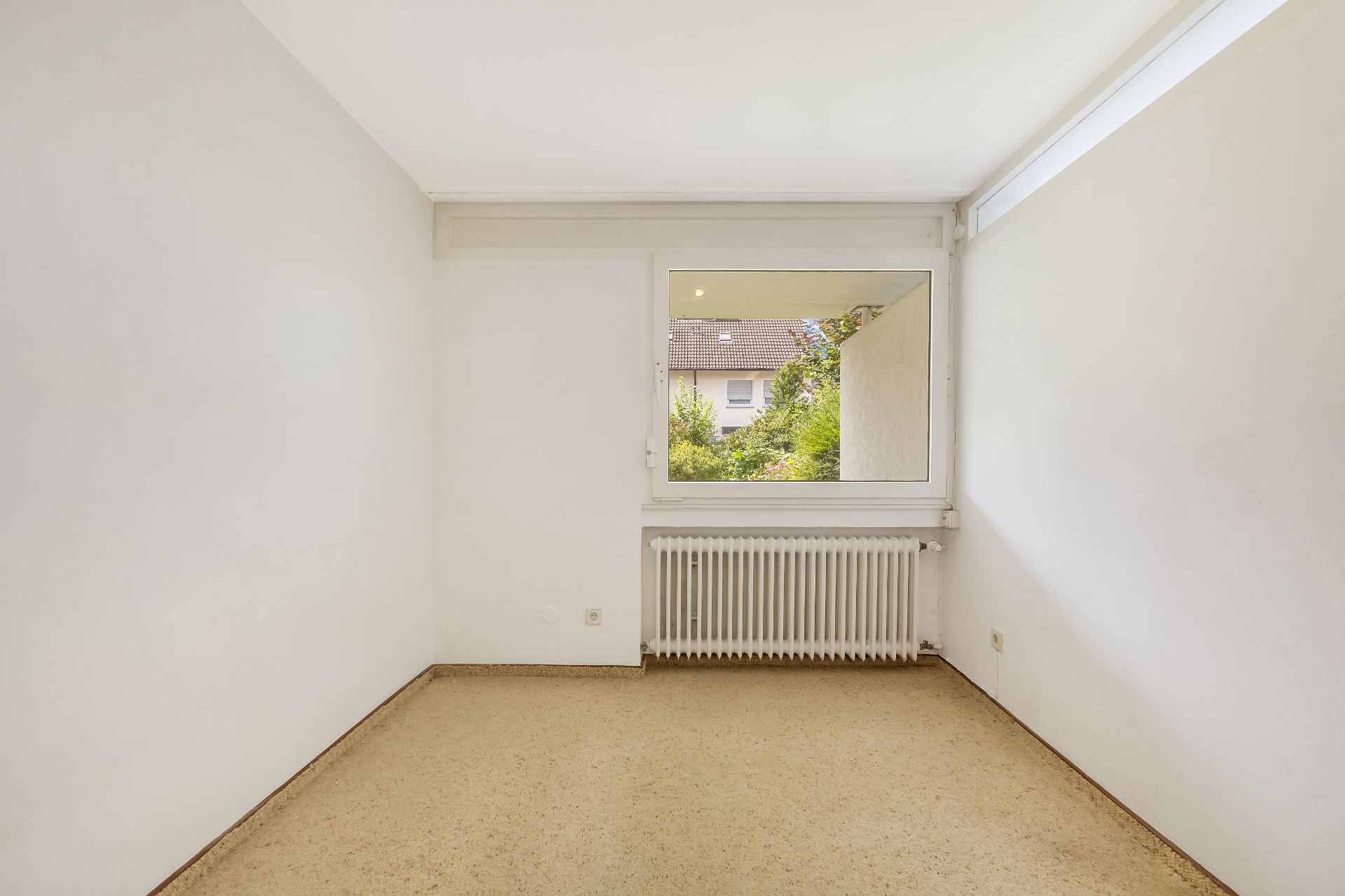 Leeres, kleines Zimmer mit beigem Boden, weißem Heizkörper und Fenster zum Garten.
