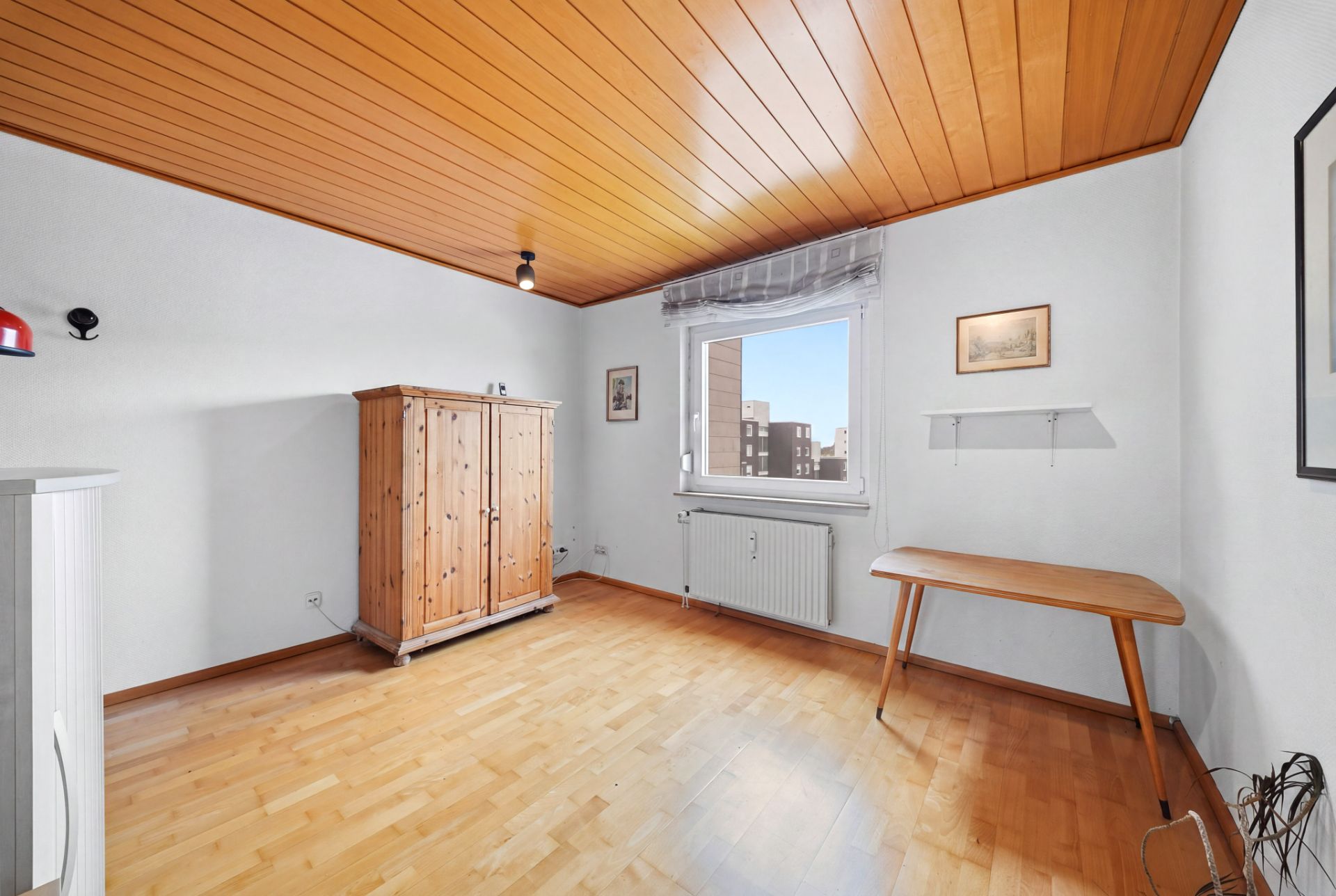 Helles Zimmer mit Holzdecke, Holzschrank, Tisch, Fenster und Bildern an weißen Wänden.