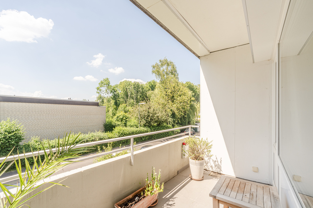 Heller Balkon mit Pflanzen, Holztisch und Blick auf Bäume und einen Nachbarbau.