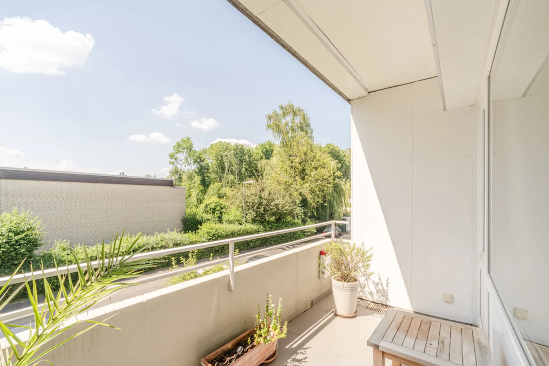 Heller Balkon mit Pflanzen, Holztisch und Blick auf Bäume und einen Nachbarbau.