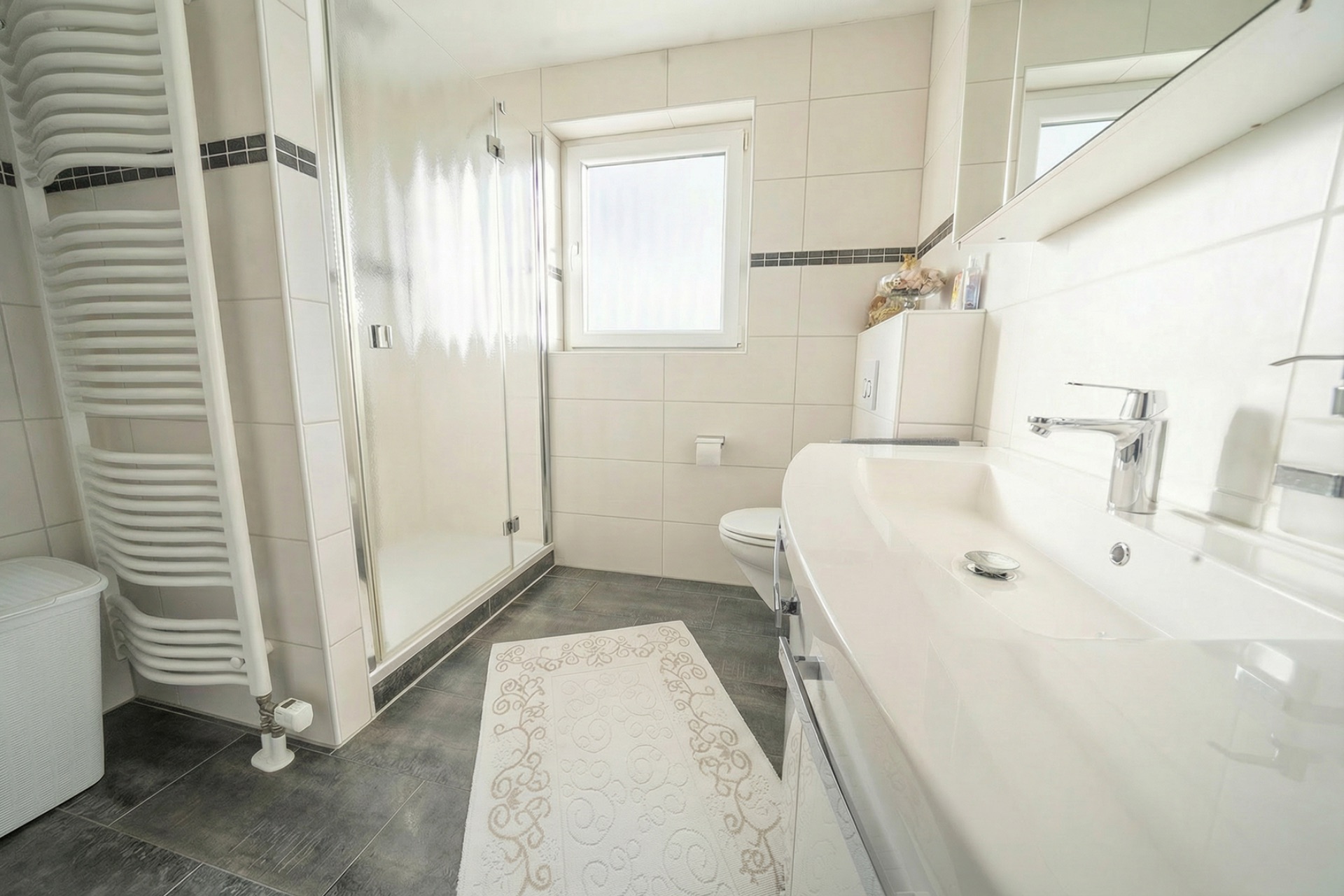Helles Badezimmer mit Dusche, Waschbecken, Spiegel und Handtuchheizkörper.