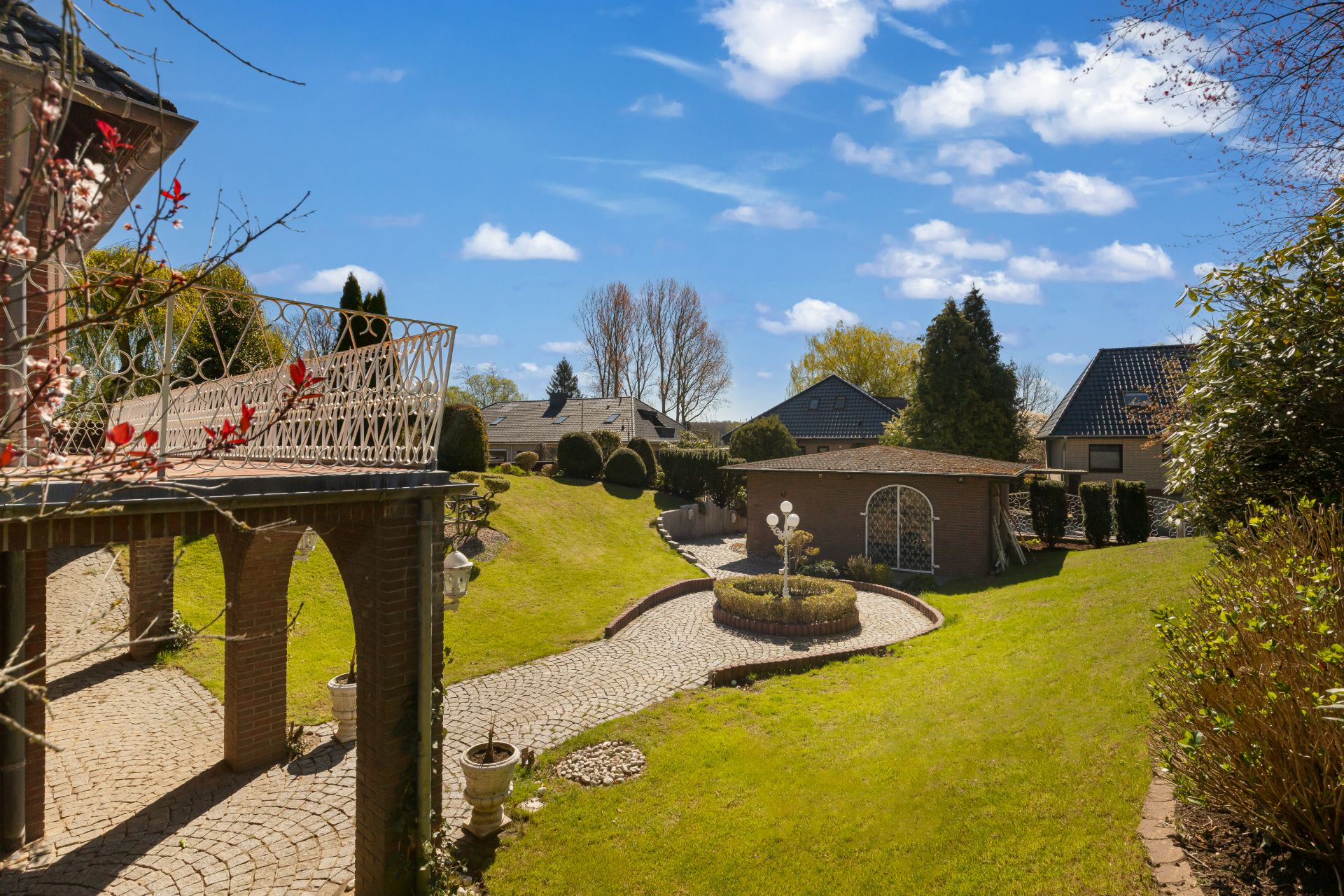 Garten mit Rasenfläche, gepflastertem Weg, Brunnen und Gartenhaus unter blauem Himmel.