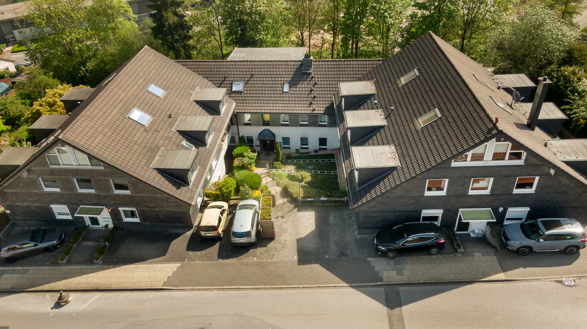 Luftaufnahme eines großen Wohngebäudes mit mehreren Autos vor dem Haus und begrüntem Eingangsbereich.