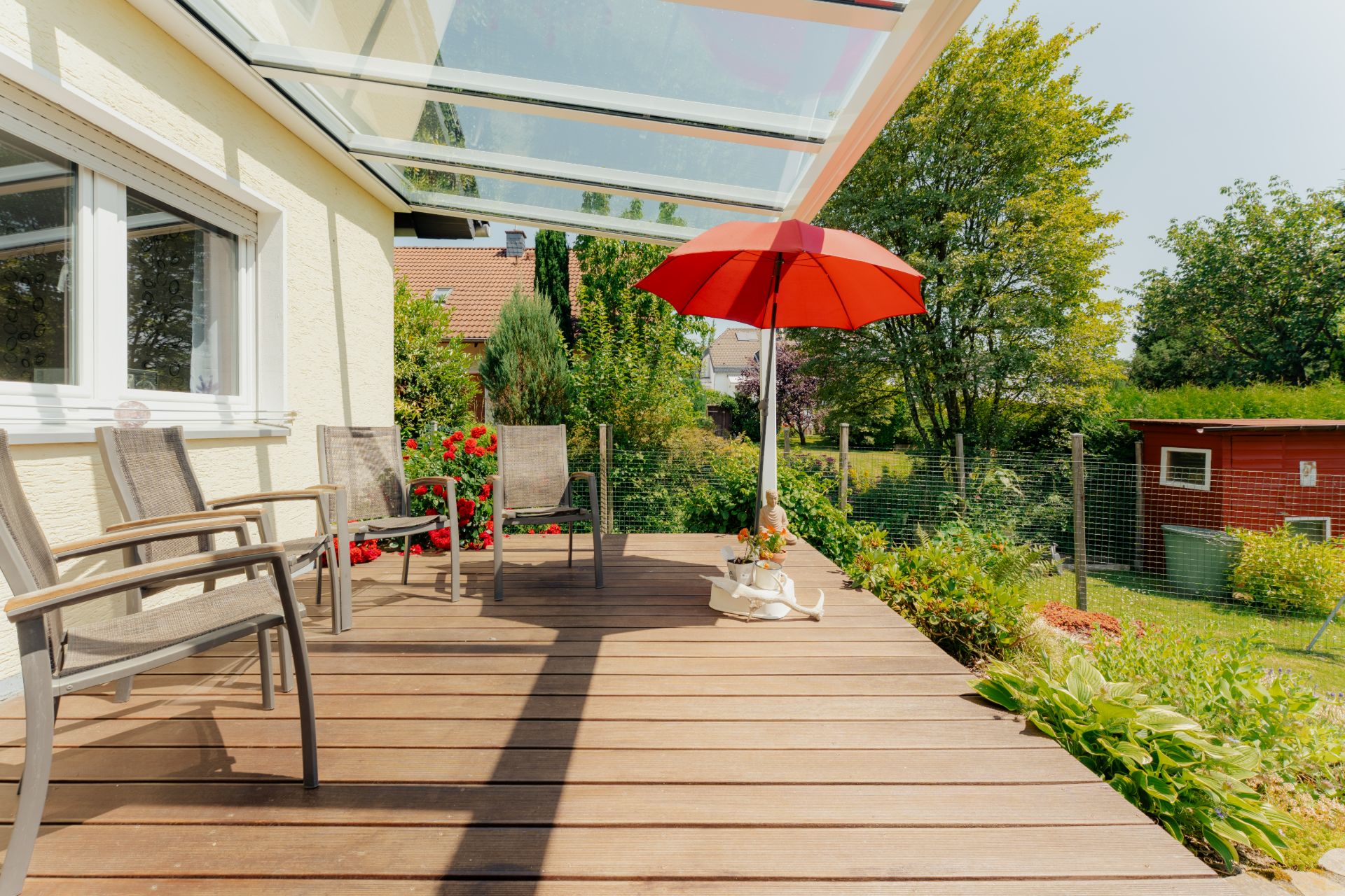 Holzterrasse mit Glasdach, mehreren Stühlen und rotem Sonnenschirm im Garten.