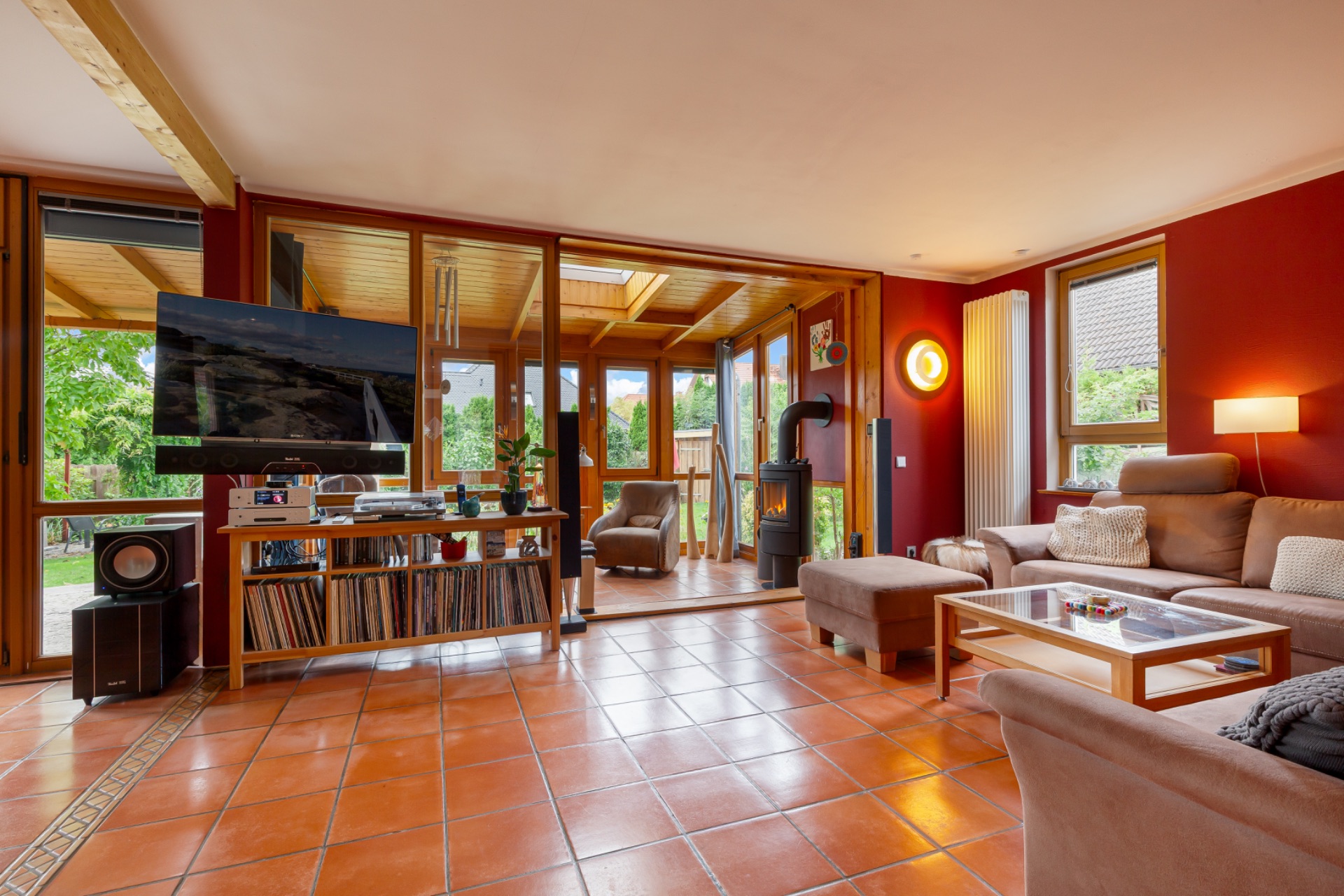 Wohnzimmer mit terrakottafarbenem Fliesenboden, Ecksofa, TV, Bücherregal und Zugang zu einem Wintergarten.