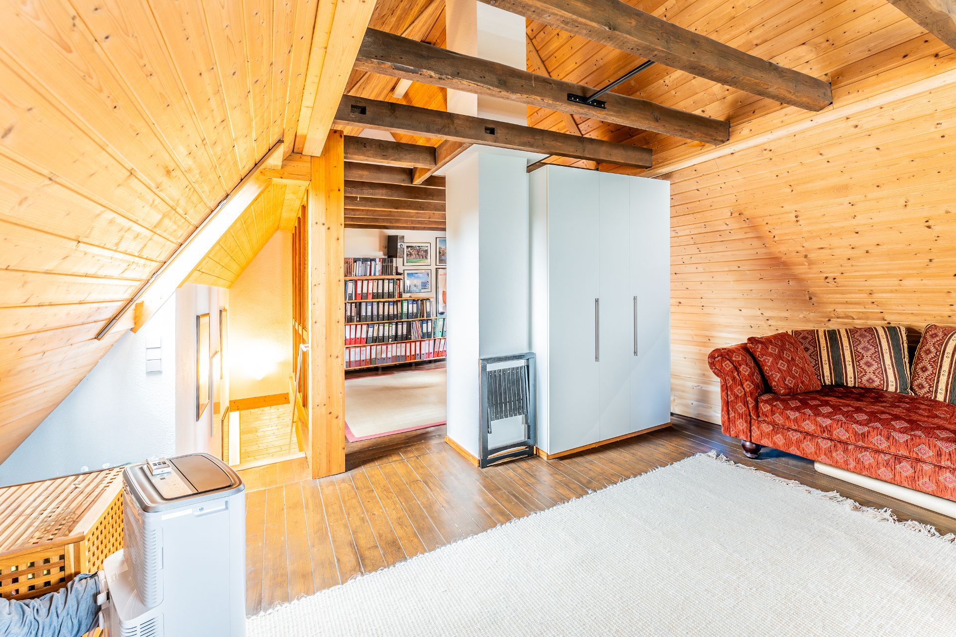 Wohnzimmer im Dachgeschoss mit Holzdecke, rotem Sofa, weißem Teppich und Schrank.