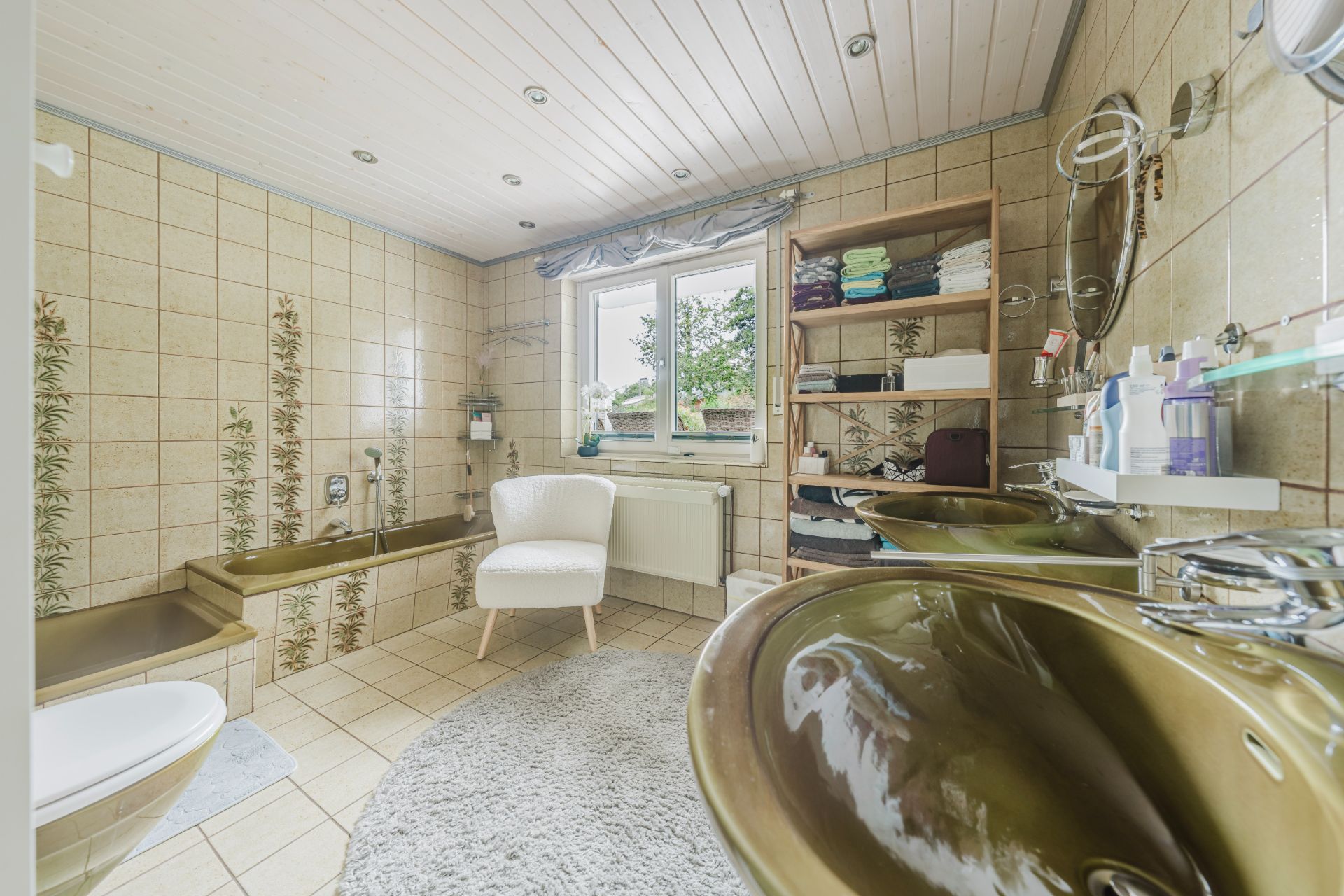 Helles Badezimmer mit zwei Waschtischen, Badewanne, Regal, Sessel und Fenster mit Blick nach draußen.