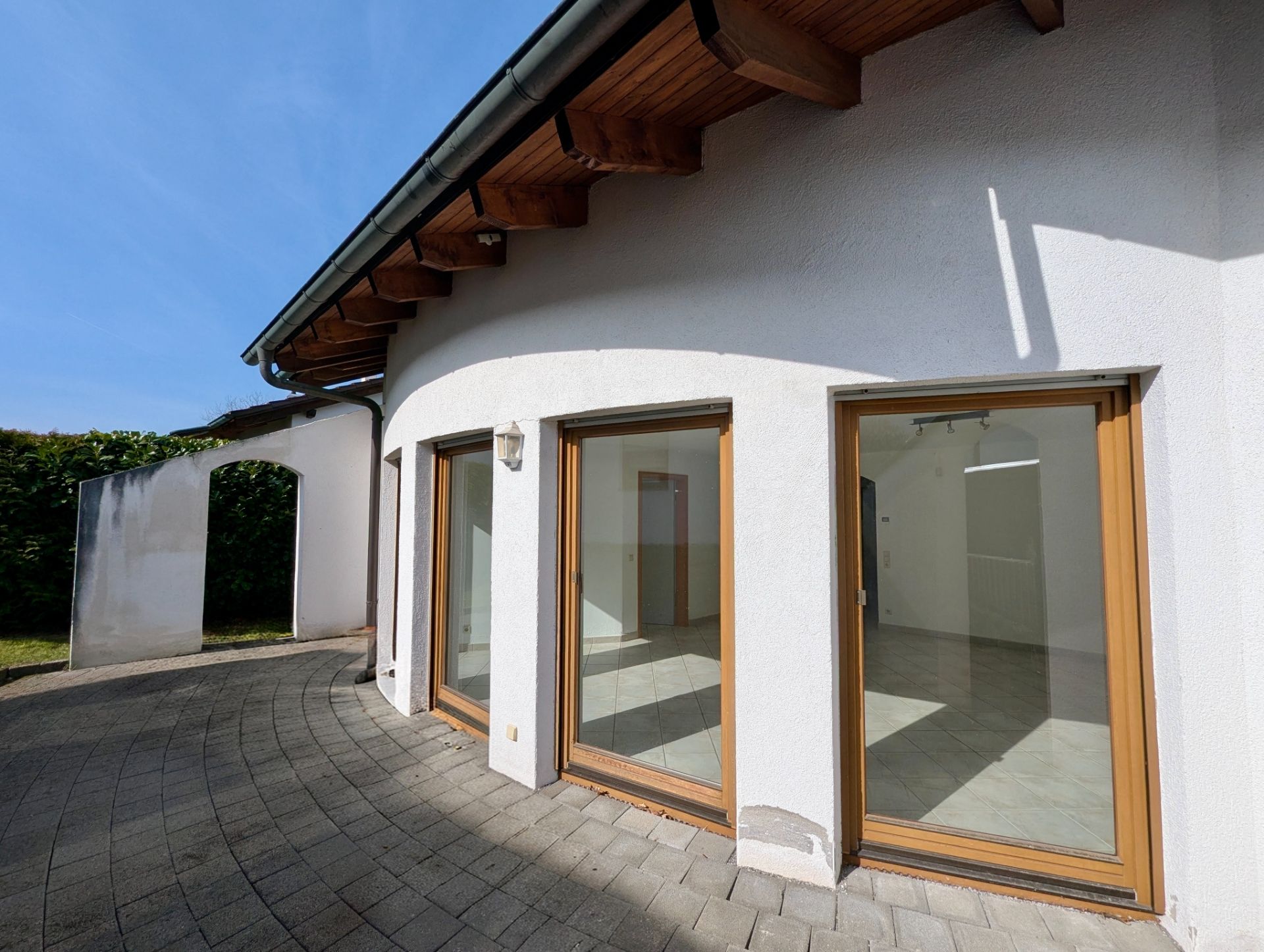 Gebogenes Haus mit Holzfenstern und gepflasterter Terrasse, große Glasfronten.