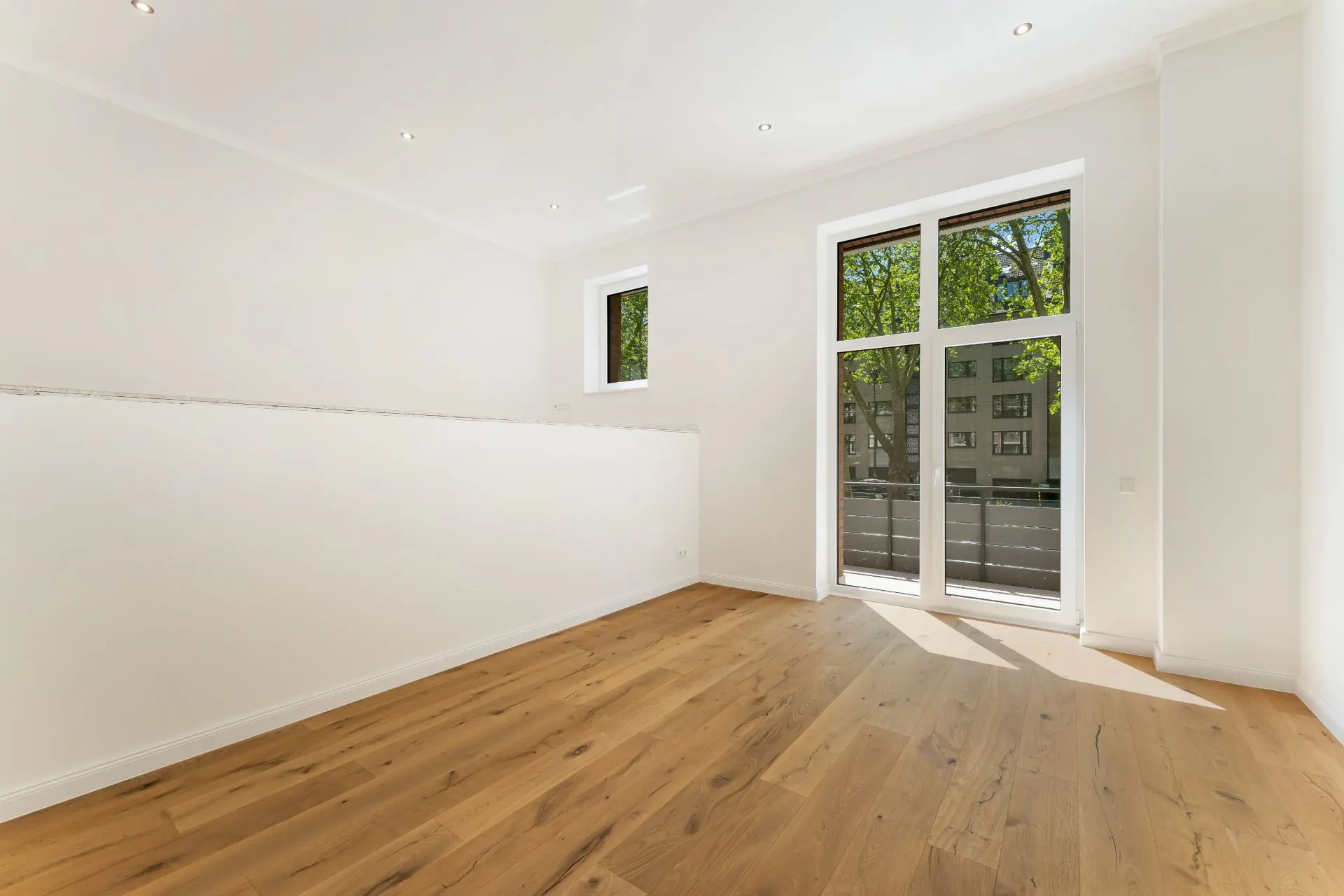 Heller Raum mit Holzboden, weißen Wänden, großem Fenster und Zugang zum Balkon.