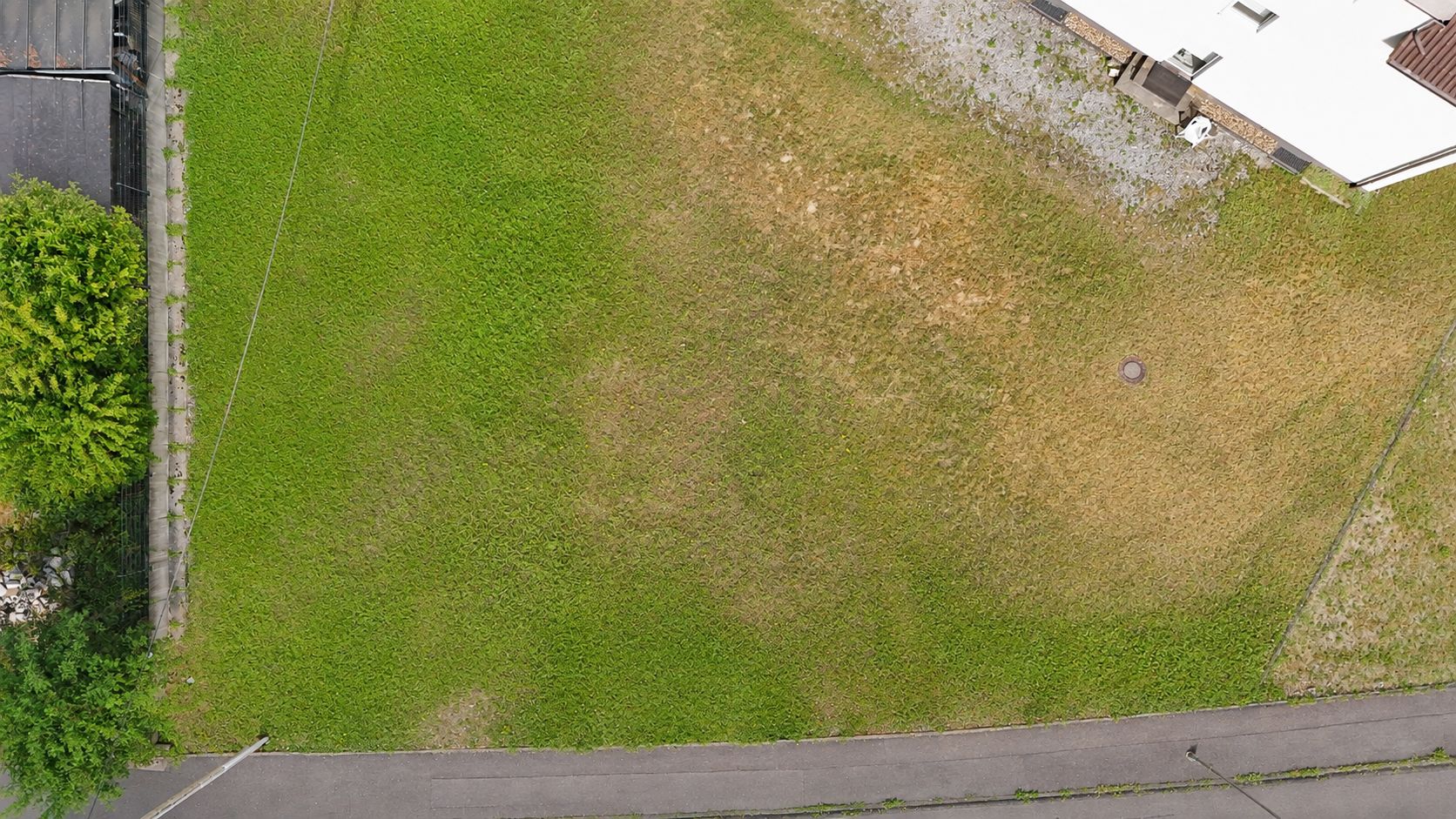 Luftaufnahme eines rechteckigen, teilweise grünen Rasens mit angrenzendem Haus und Gehweg.