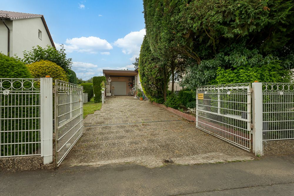 Offenes Einfahrtstor mit gepflasterter Zufahrt zu einem Haus und Garage, umgeben von Büschen.