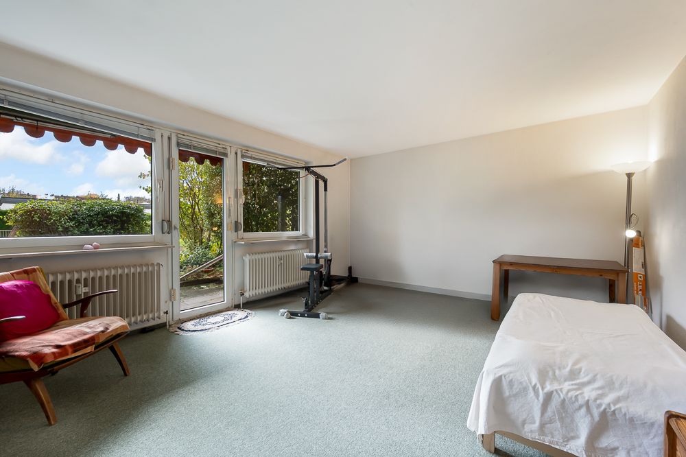 Helles Zimmer mit einem Bett, einem Tisch, einem Sessel und Fitnessgerät, große Fenster mit Blick nach draußen.