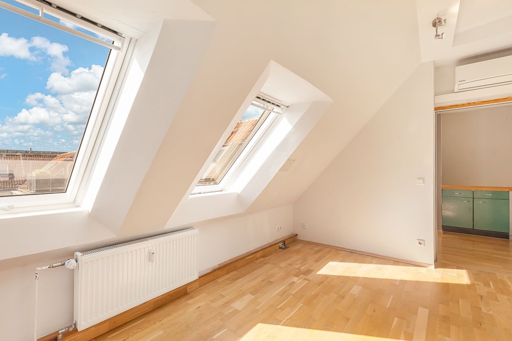 Helles Zimmer mit Dachschrägen, zwei großen Fenstern und Parkettboden.