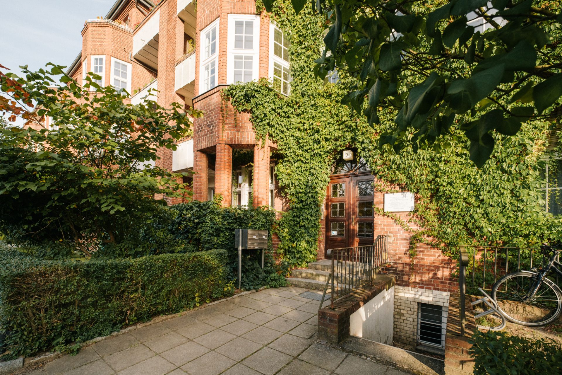 Backsteinwohnhaus mit Efeu an der Fassade, Eingangstür, Fahrrad und viel Grün.