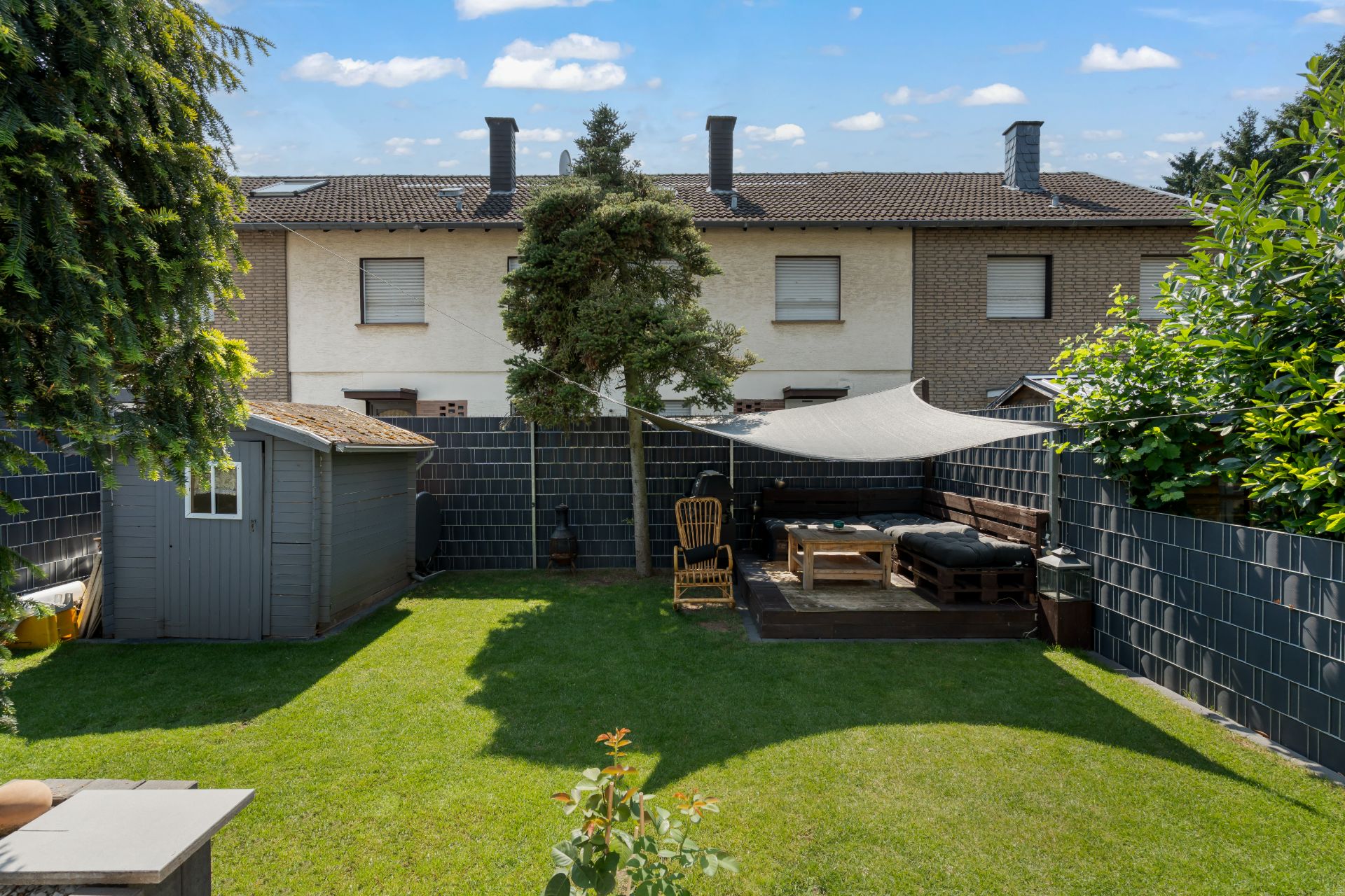 Garten mit Rasen, Holzterrasse, Sitzbereich, Gartenhaus und Baum, von Zaun umgeben.