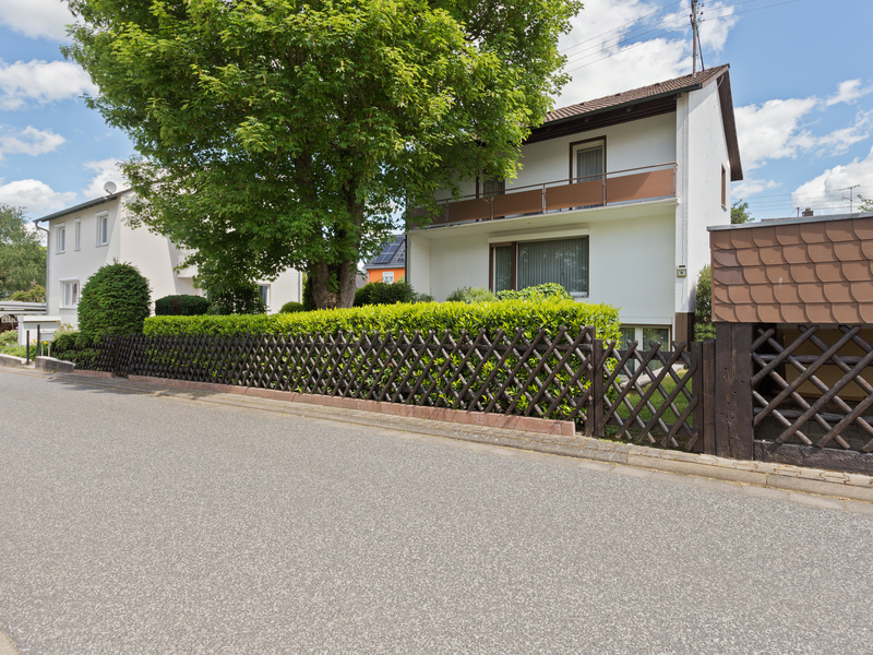 Zweistöckiges Wohnhaus mit Garten, Holzzaun und großem Baum an einer ruhigen Straße.