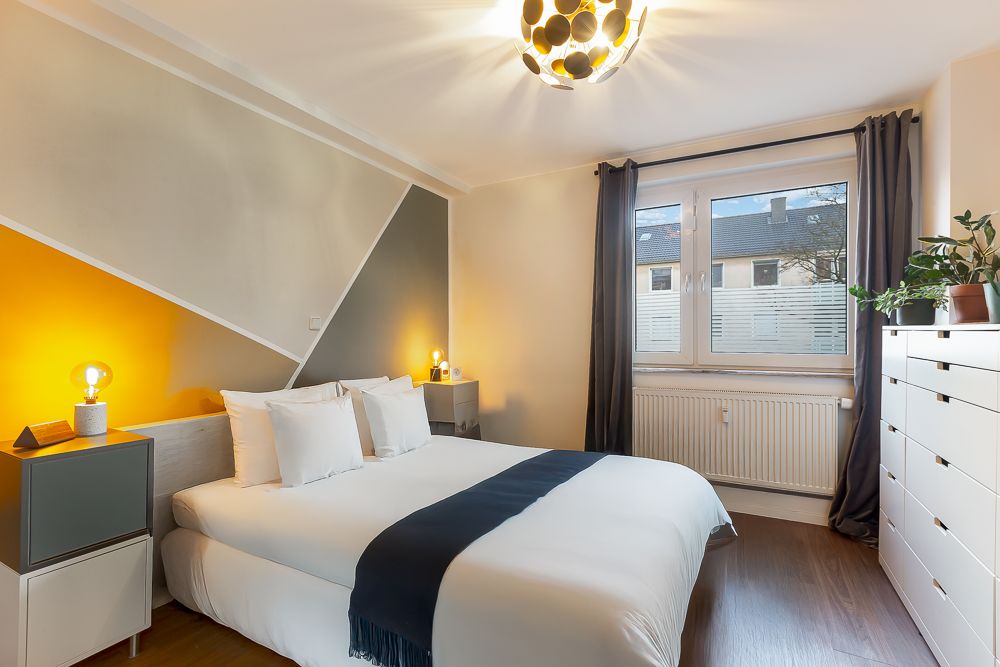 Modernes Schlafzimmer mit Doppelbett, Nachttischen, Kommode und großem Fenster mit Vorhängen.