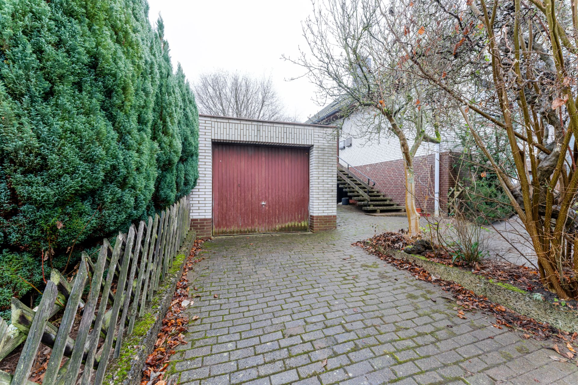 Gepflasterte Einfahrt mit Garage, Holzzaun, Bäumen und außenliegender Treppe zum Haus.